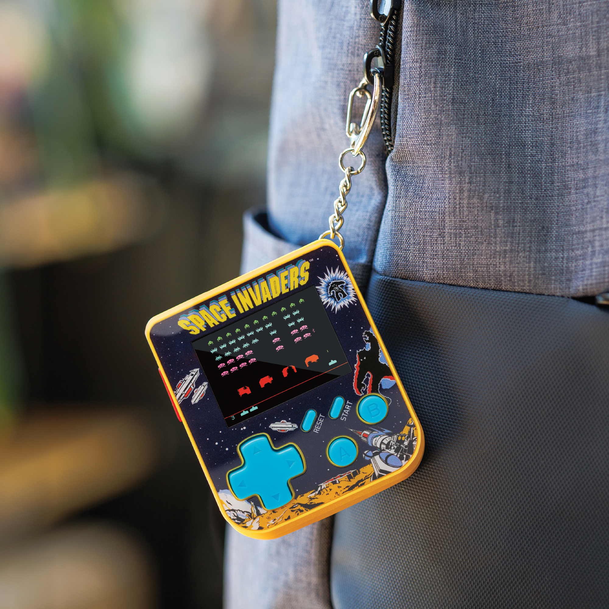 Ein Space Invaders Handheld-Spiel hängt an einer silbernen Kette. Das Spiel ist gelb, blau und schwarz. Es hängt an einem grauen Rucksack.