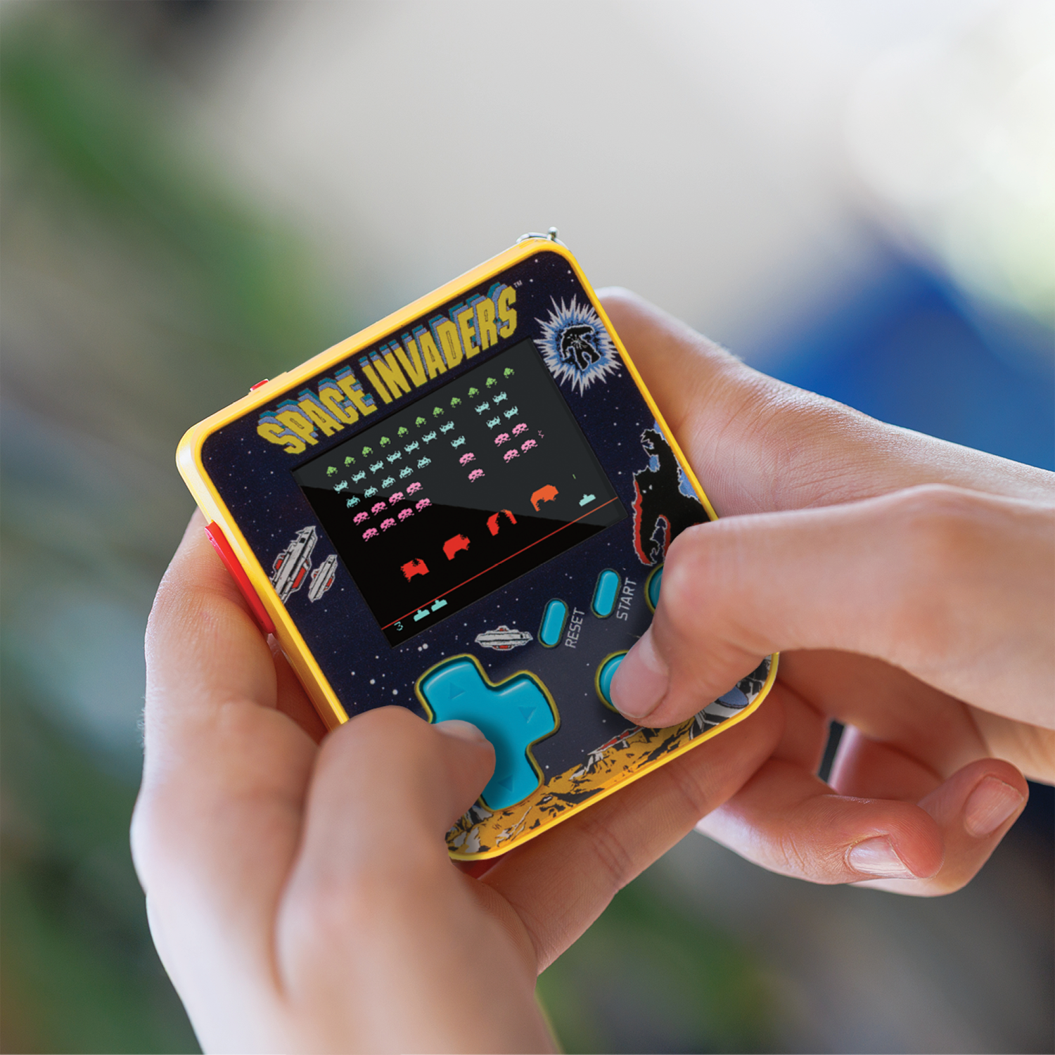 Eine Person hält ein Space Invaders-Handheld-Spiel. Der Bildschirm zeigt das Spiel. Das Spiel ist bunt und die Knöpfe sind blau.