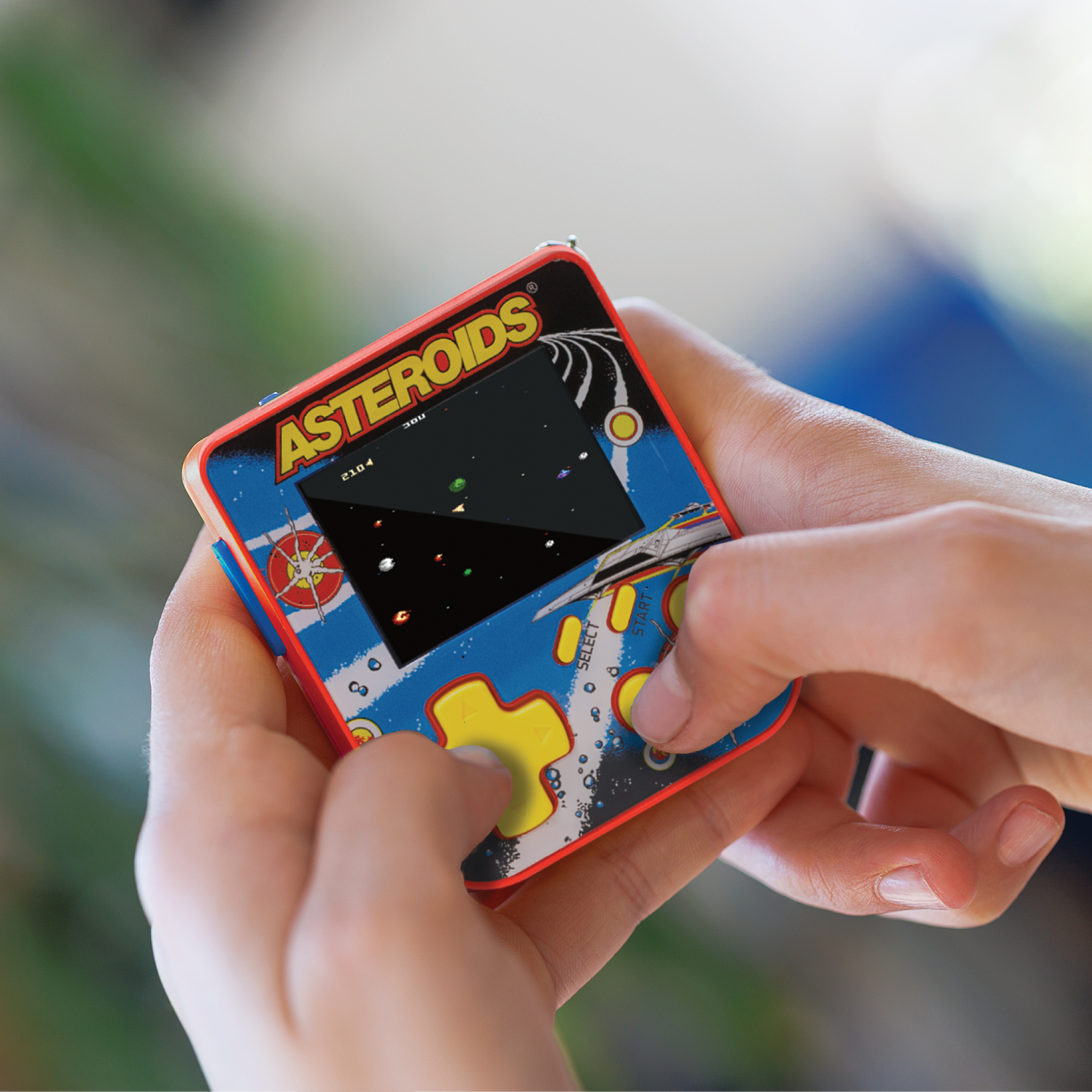 Hände halten ein Retro-Asteroids-Spiel. Der Bildschirm zeigt das Spiel. Das Spiel hat ein blaues und rotes Gehäuse, und die Knöpfe sind gelb.