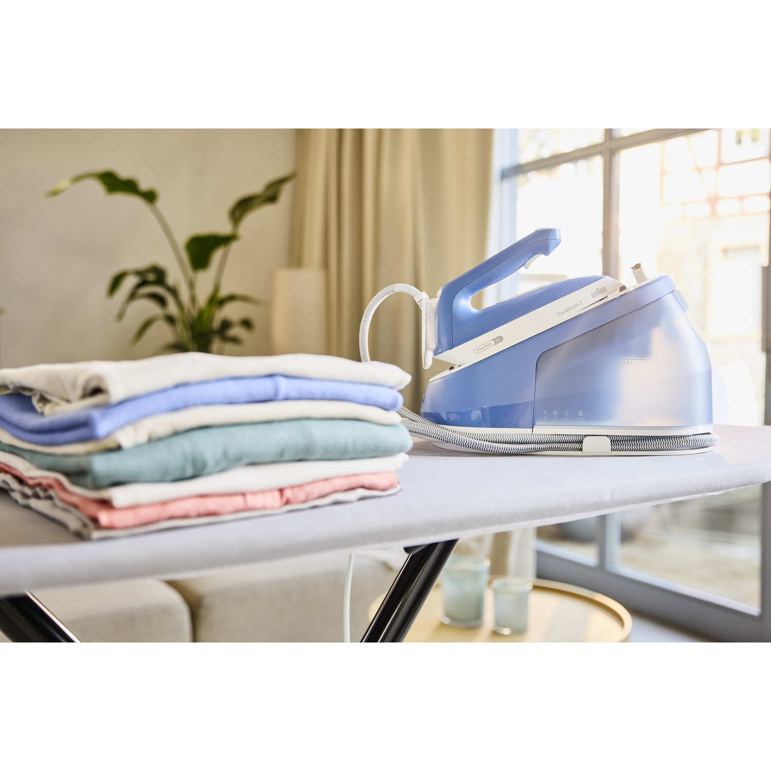 Blue iron and iron board with stack of clothes. Window and plant in the background. Clean and bright room.