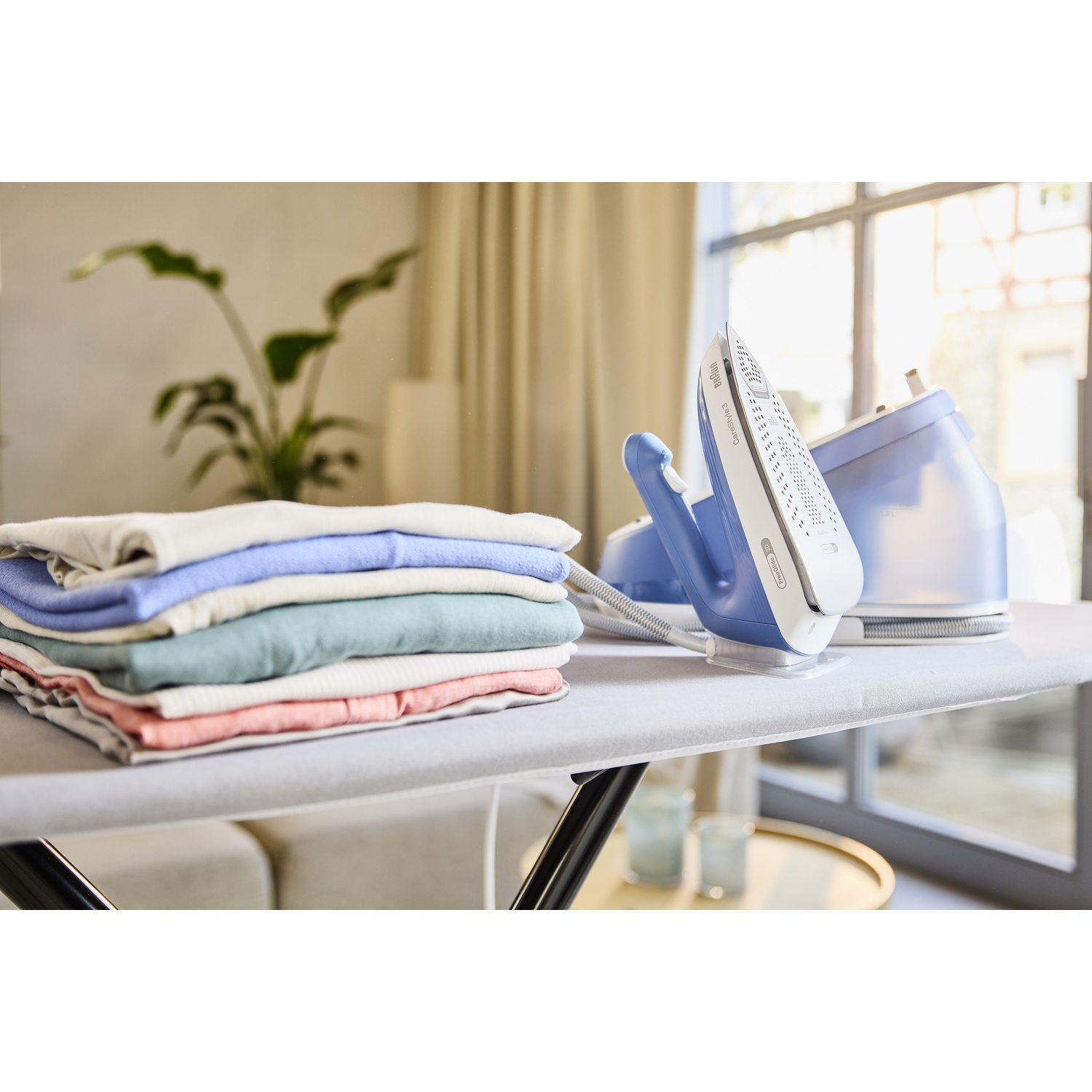 An ironing board with a blue iron and a stack of folded clothes. A plant and window are in the background. Two glasses are on the board.