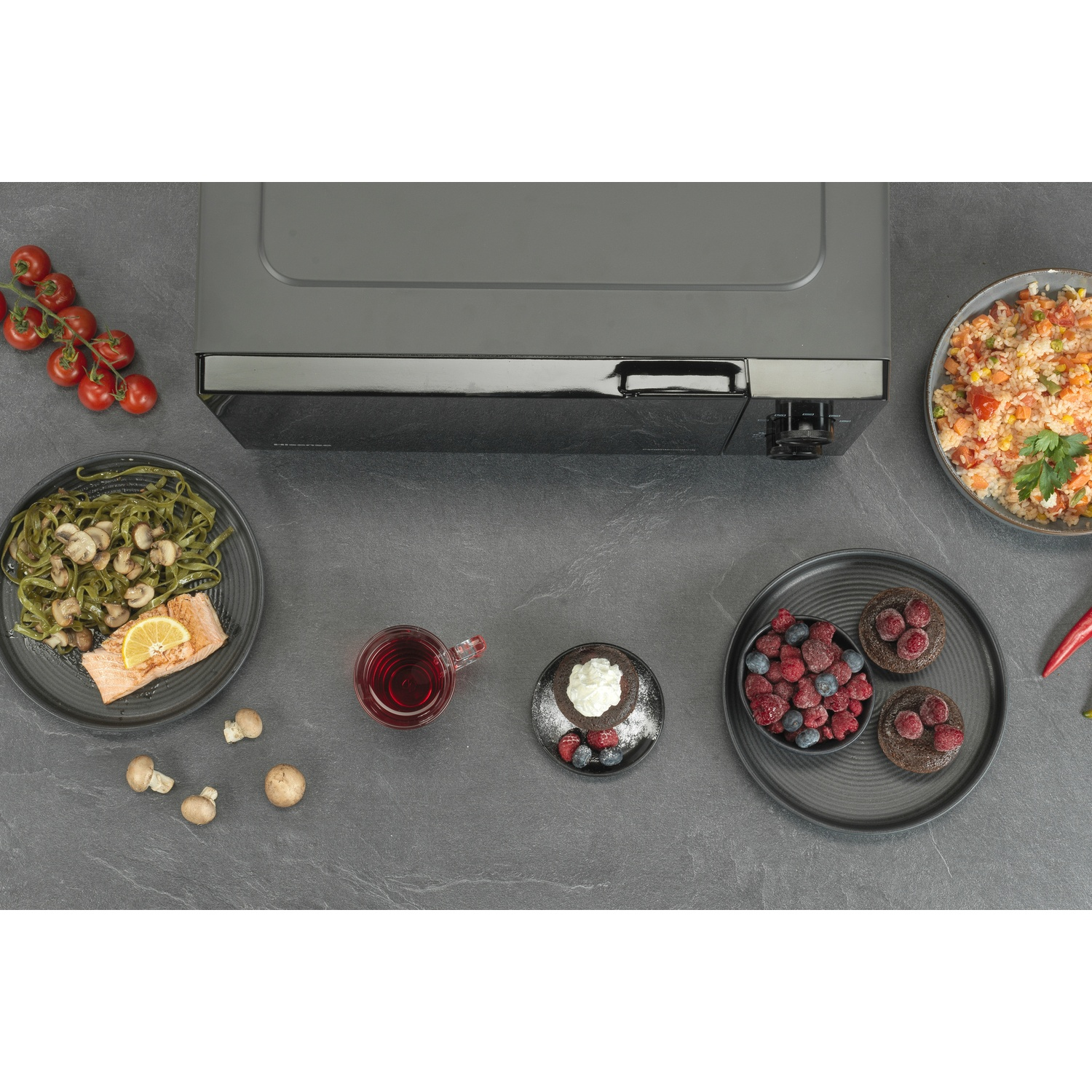 Vista dall'alto di vari piatti: pasta, riso, dessert, pomodori e un microonde su una superficie grigio scuro.