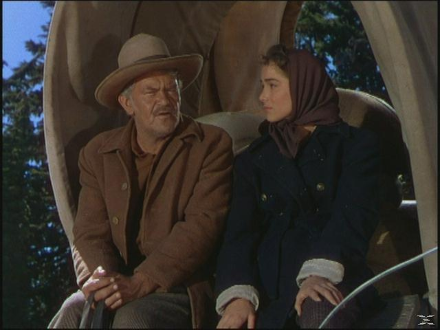 Ein älterer Mann und eine junge Frau sitzen in einem Wagen. Der Mann trägt einen Hut und einen Mantel. Die Frau trägt ein Kopftuch und einen Mantel.