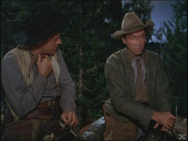 Zwei Männer in Cowboy-Kleidung sitzen im Freien. Einer trägt einen schwarzen Hut und eine Weste, der andere einen braunen Hut und eine Jacke. Wald im Hintergrund.