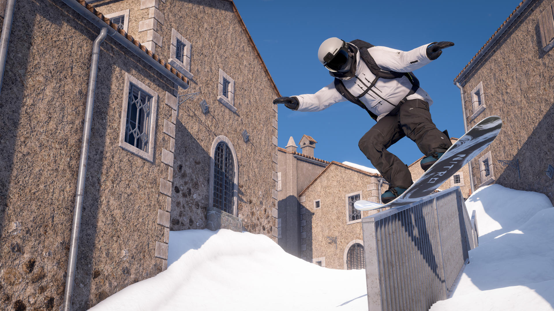 Ein Snowboarder in weißer Ausrüstung springt über eine schneebedeckte Leiste vor dem Hintergrund von Steingebäuden unter blauem Himmel.