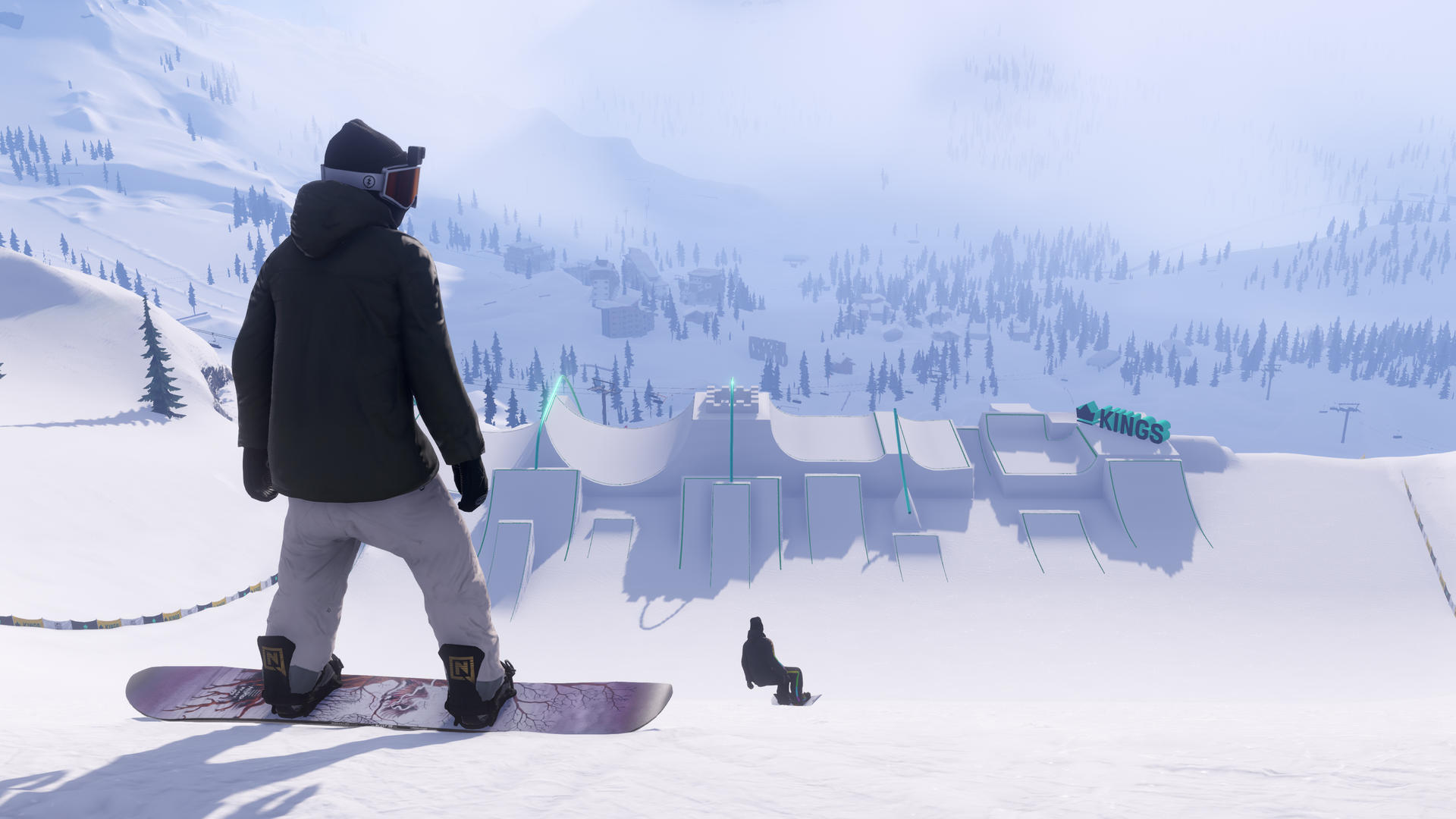 Ein Snowboarder steht auf einem schneebedeckten Hang und blickt auf einen Snowpark. Ein anderer Snowboarder ist in der Luft. Berge und Bäume im Hintergrund.