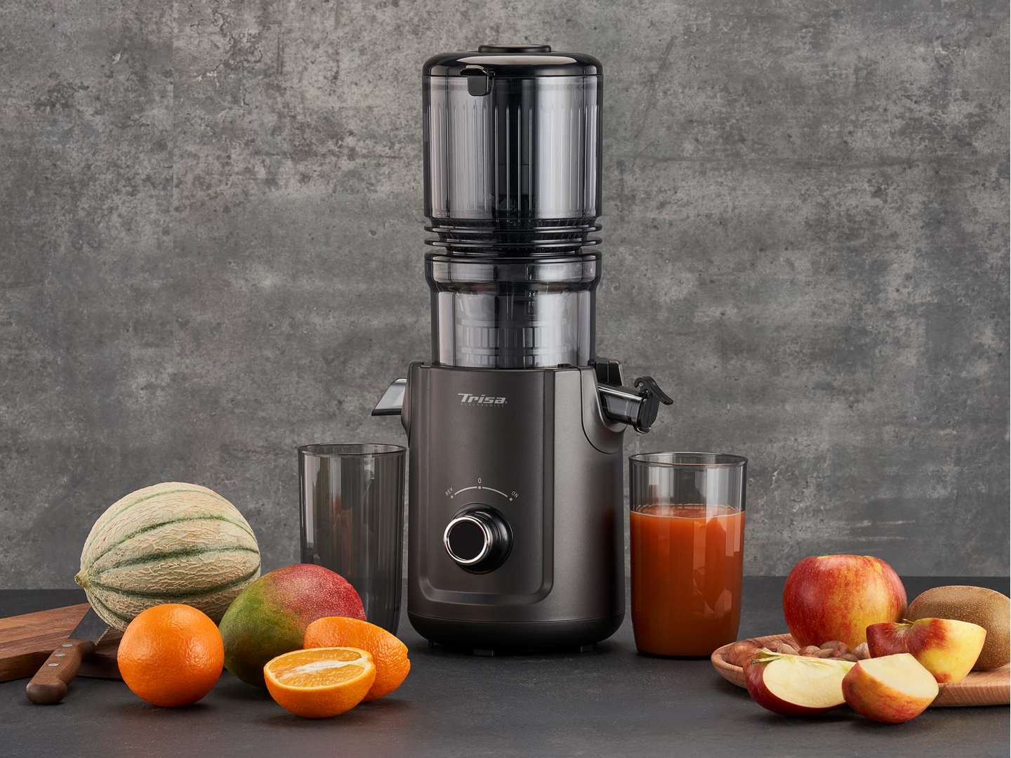 Un estrattore di succo grigio scuro con frutta, tra cui arance e mele, e un bicchiere di succo. Un melone a sinistra.