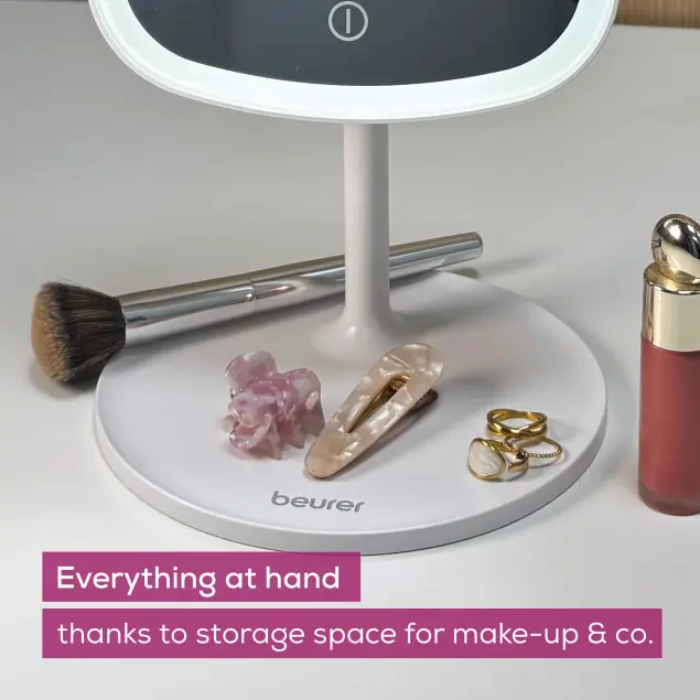 Ein weißer Schminkspiegel mit Zubehör. Auf der Basis befinden sich ein Pinsel, Haarspangen, Ringe und eine Make-up-Flasche.