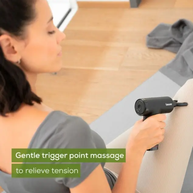 Eine Frau benutzt ein schwarzes Massagegerät an ihrem Bein. Sie sitzt auf einer Matte, trägt ein graues Hemd. Der Hintergrund ist ein Holzboden.
