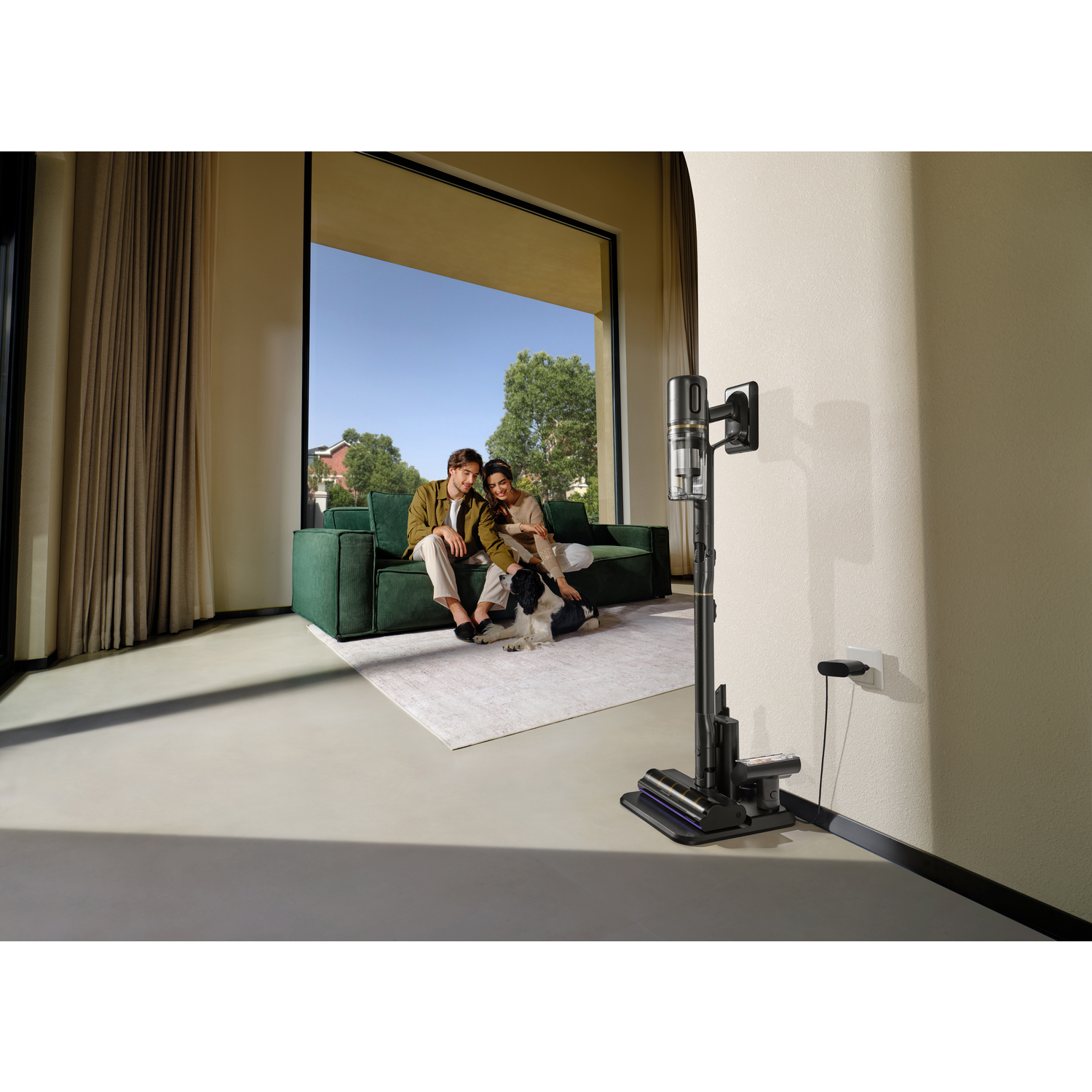 A couple and a dog sit on a green sofa. A vacuum cleaner is charging nearby. A large window overlooks a sunny outdoor scene.