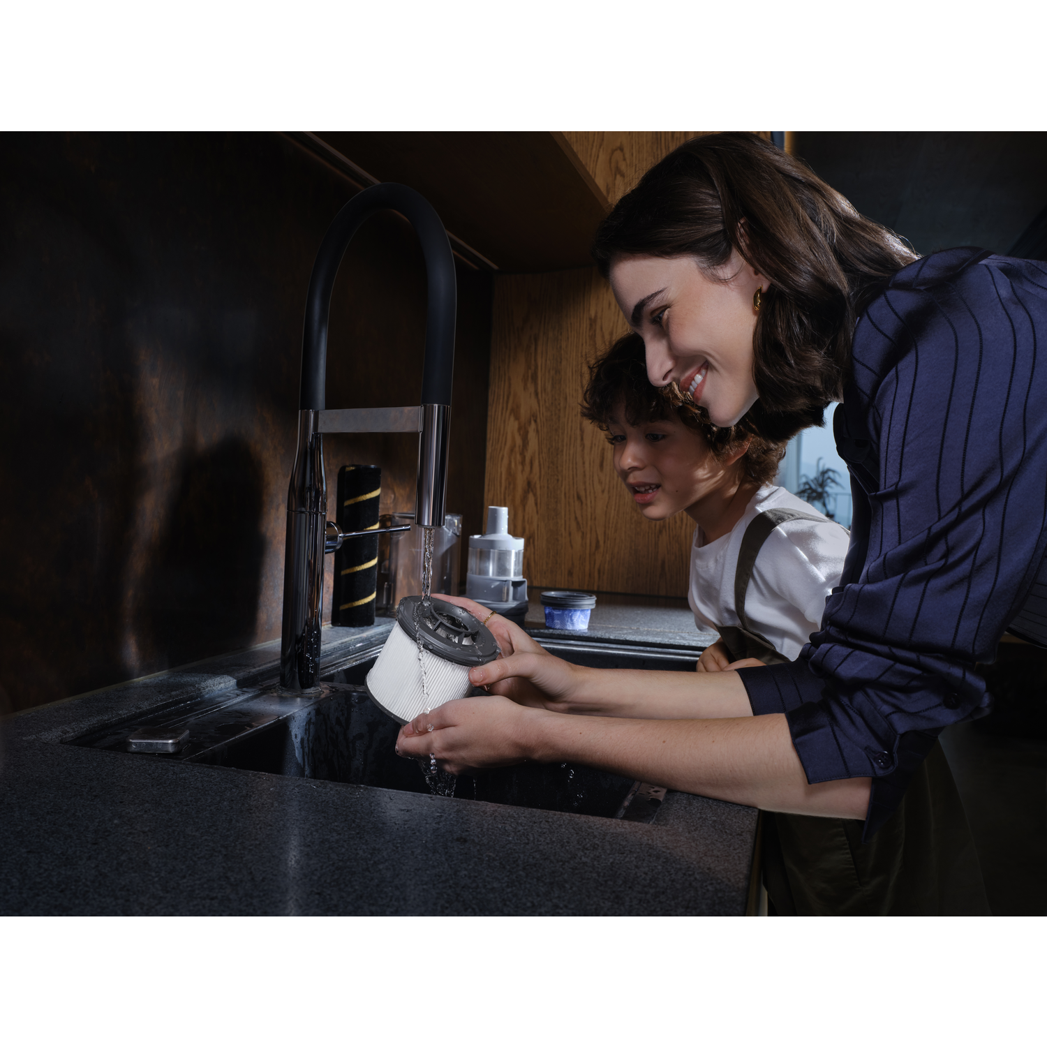 A woman and child wash a vacuum filter at a sink. Water runs over the filter. The woman smiles. Dark cabinets and sink in the background.