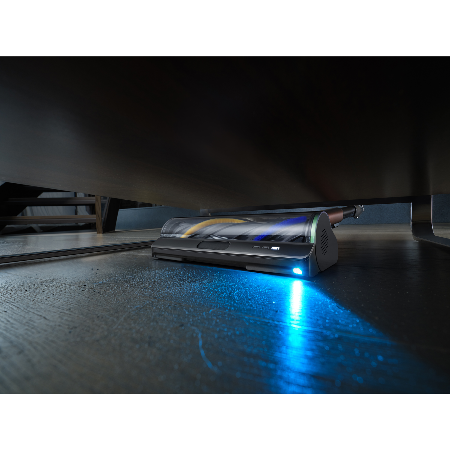 A vacuum cleaner is placed under a dark brown table. The floor is dark, and the cleaner has a blue light on. 