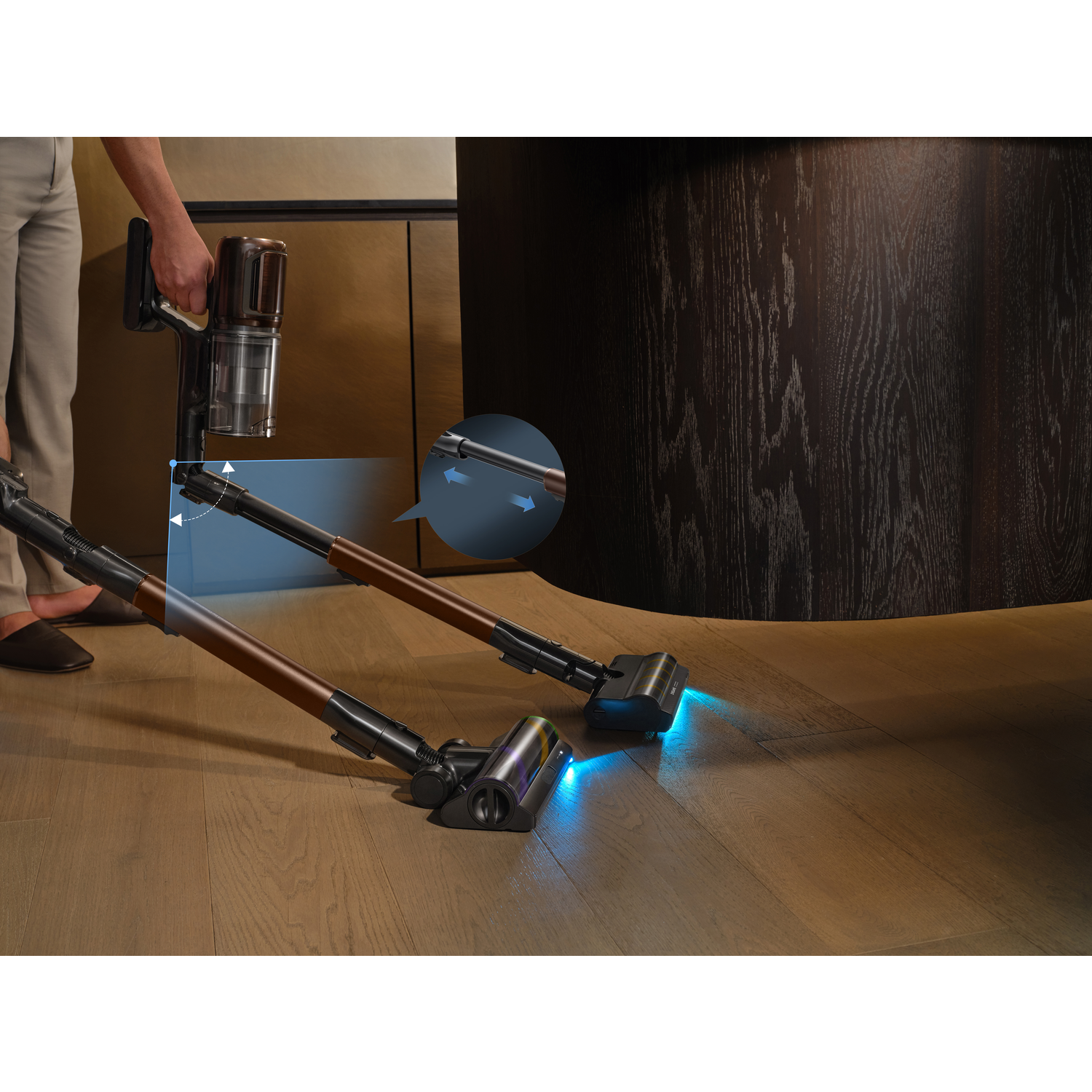 A person holds a brown and black vacuum cleaner. Another vacuum cleaner is on the wooden floor, with blue lights on.