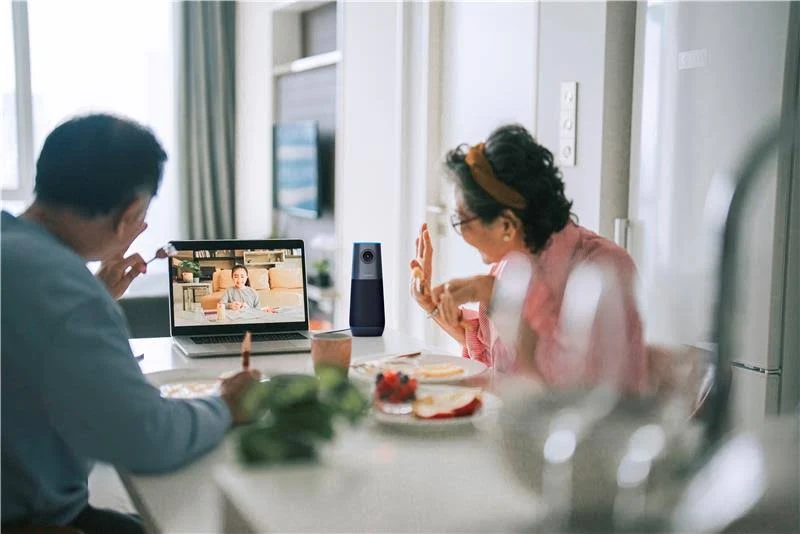 Dos personas están en una videollamada. Una está comiendo y otra saludando. Una cámara está en la mesa y una laptop está abierta.