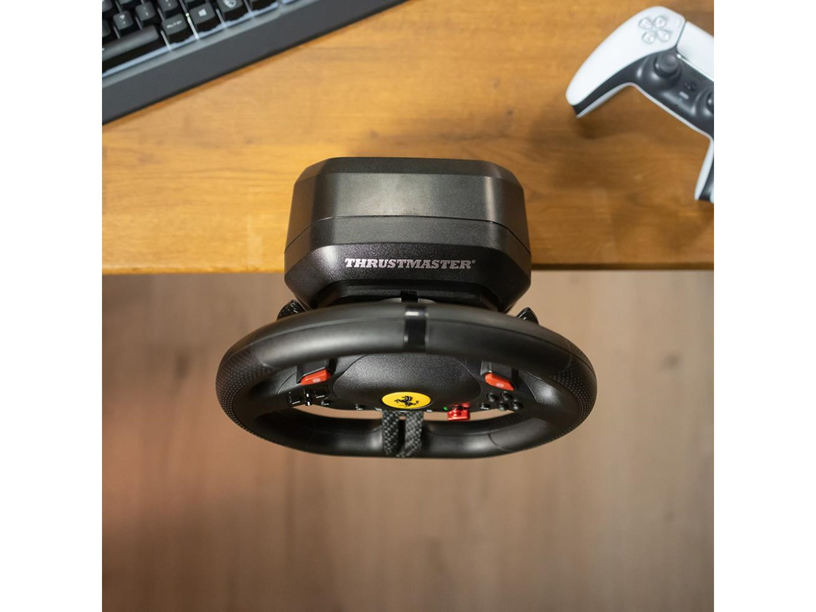 Volante de carreras Thrustmaster negro con botones rojos. Un mando blanco de PS5 está a su lado. También se ve un teclado.