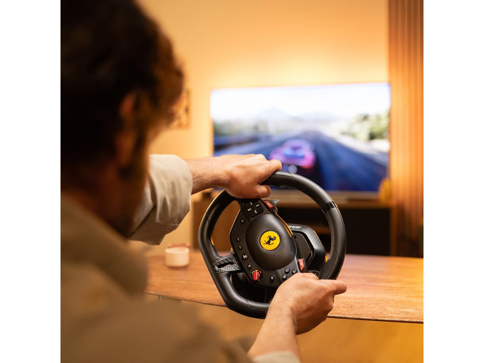 Una persona sostiene un volante Ferrari, jugando un juego de carreras en la TV. La habitación tiene iluminación cálida y una mesa de madera.