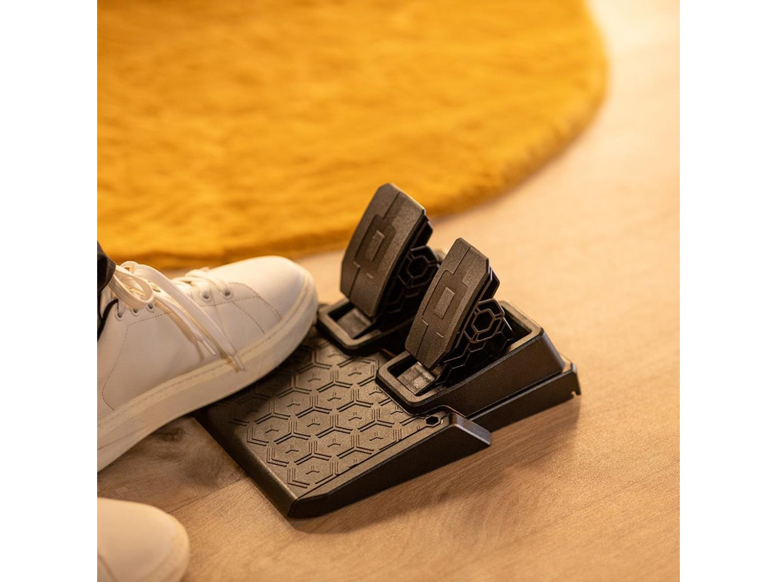 Pedales negros con un zapato. El zapato es blanco con cordones. El fondo muestra una alfombra amarilla y suelo de madera.