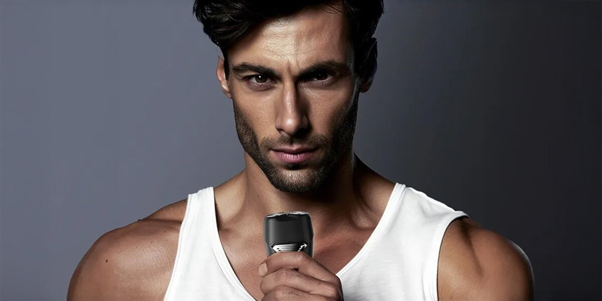 Mężczyzna w białym podkoszulku trzyma czarną golarkę elektryczną. Patrzy w kamerę. Szare tło.