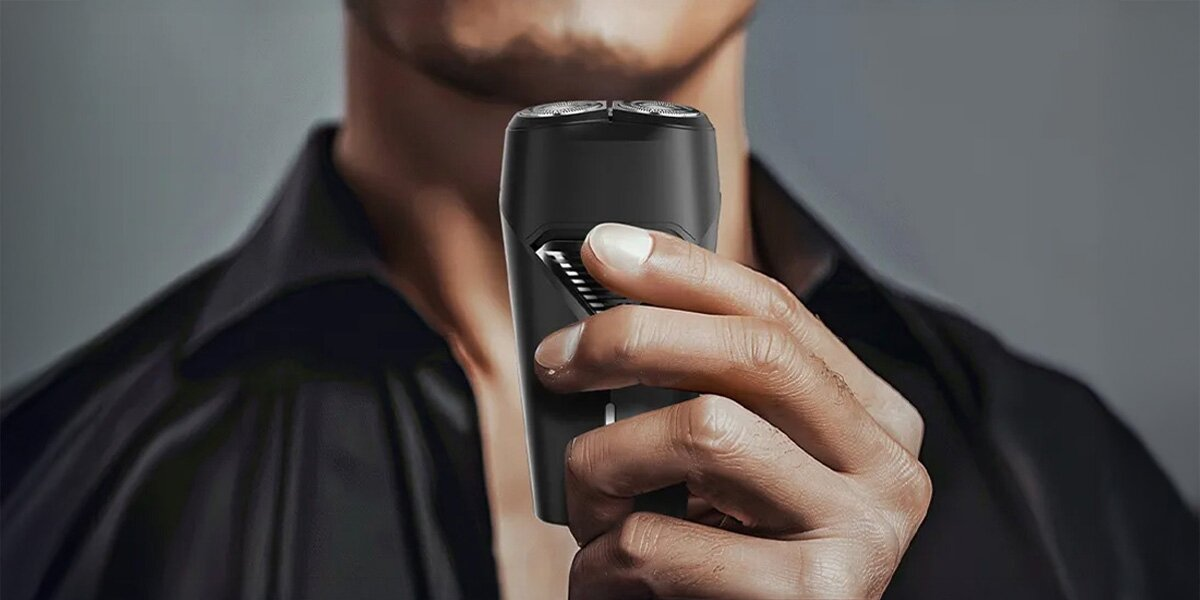 Mężczyzna trzyma czarną golarkę elektryczną. Ma na sobie czarną koszulę na szarym tle. Golarka jest w centrum.