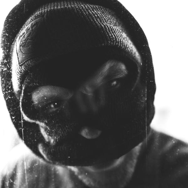 Eine Person trägt eine schwarze Skimaske und Mütze. Der Fokus liegt auf den Augen. Der Hintergrund ist weiß mit körniger Textur.