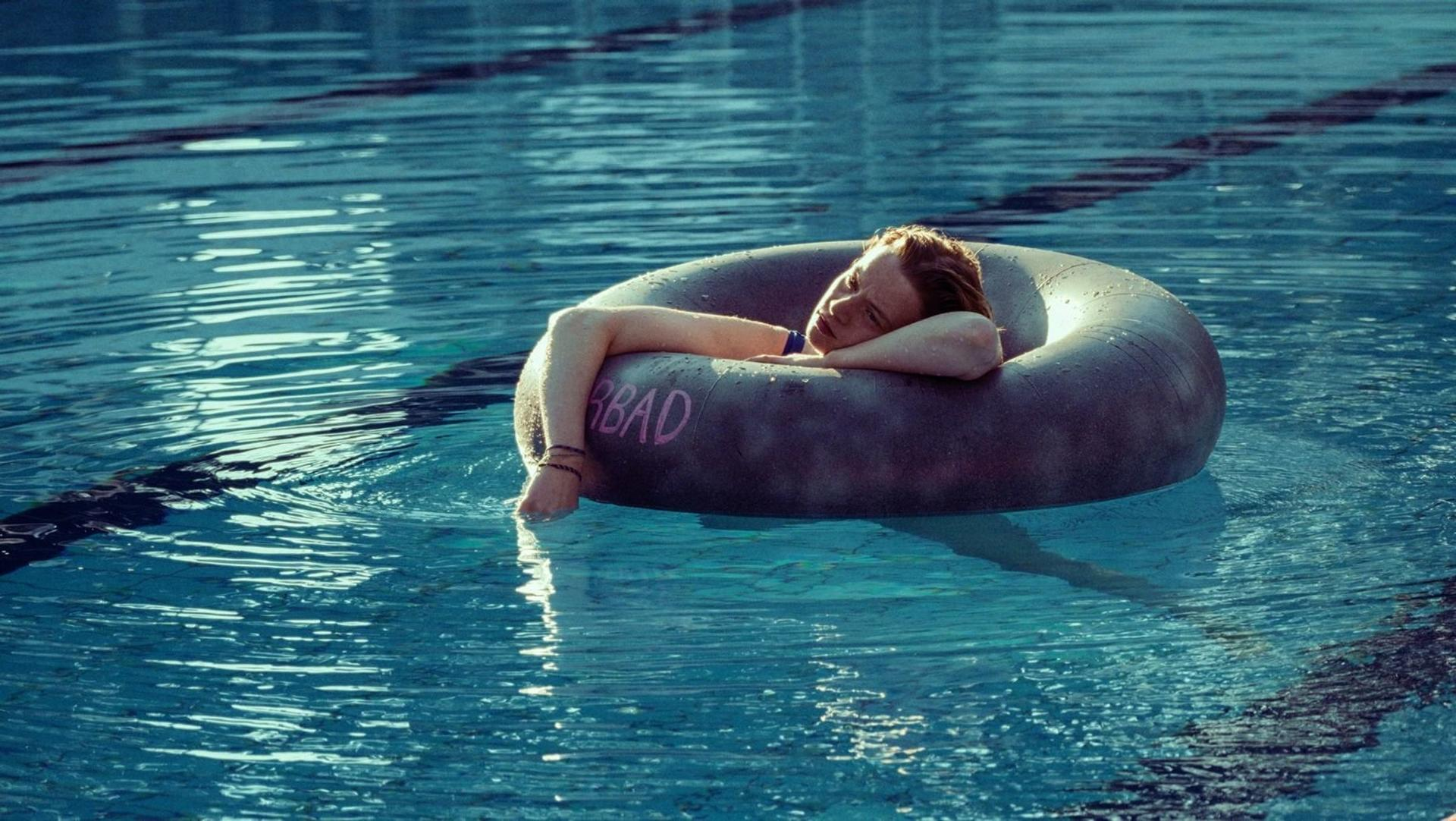 Eine Person schwimmt in einem Pool auf einem dunklen Reifen. Die Person stützt den Kopf auf die Röhre, das Wort 'BAD' ist sichtbar.