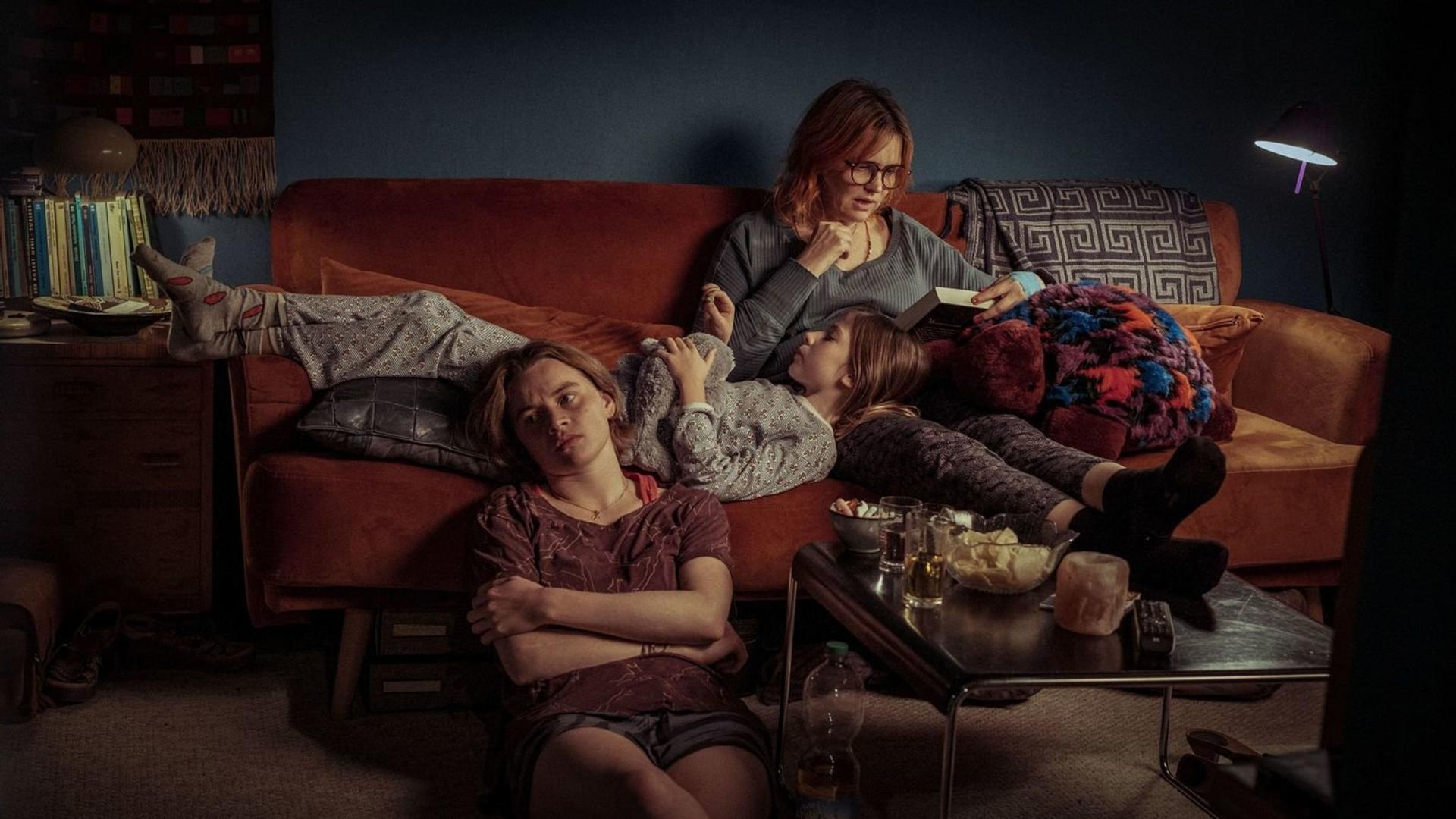 Drei Personen auf einem Sofa. Einer liest, zwei entspannen sich. Tisch mit Snacks. Mädchen auf dem Boden.