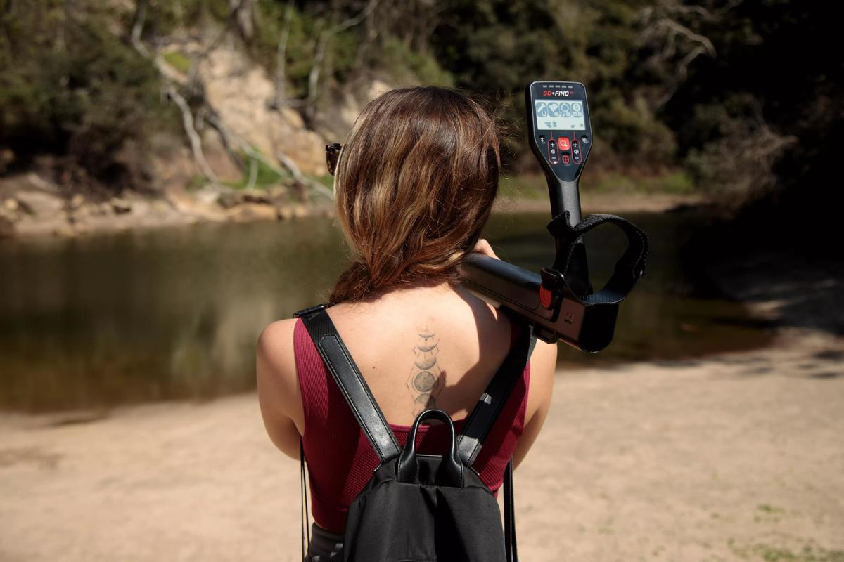 Frau mit Metalldetektor am Strand. Fluss und Bäume im Hintergrund. Tattoo auf dem Rücken. Sie hält einen schwarzen Metalldetektor.