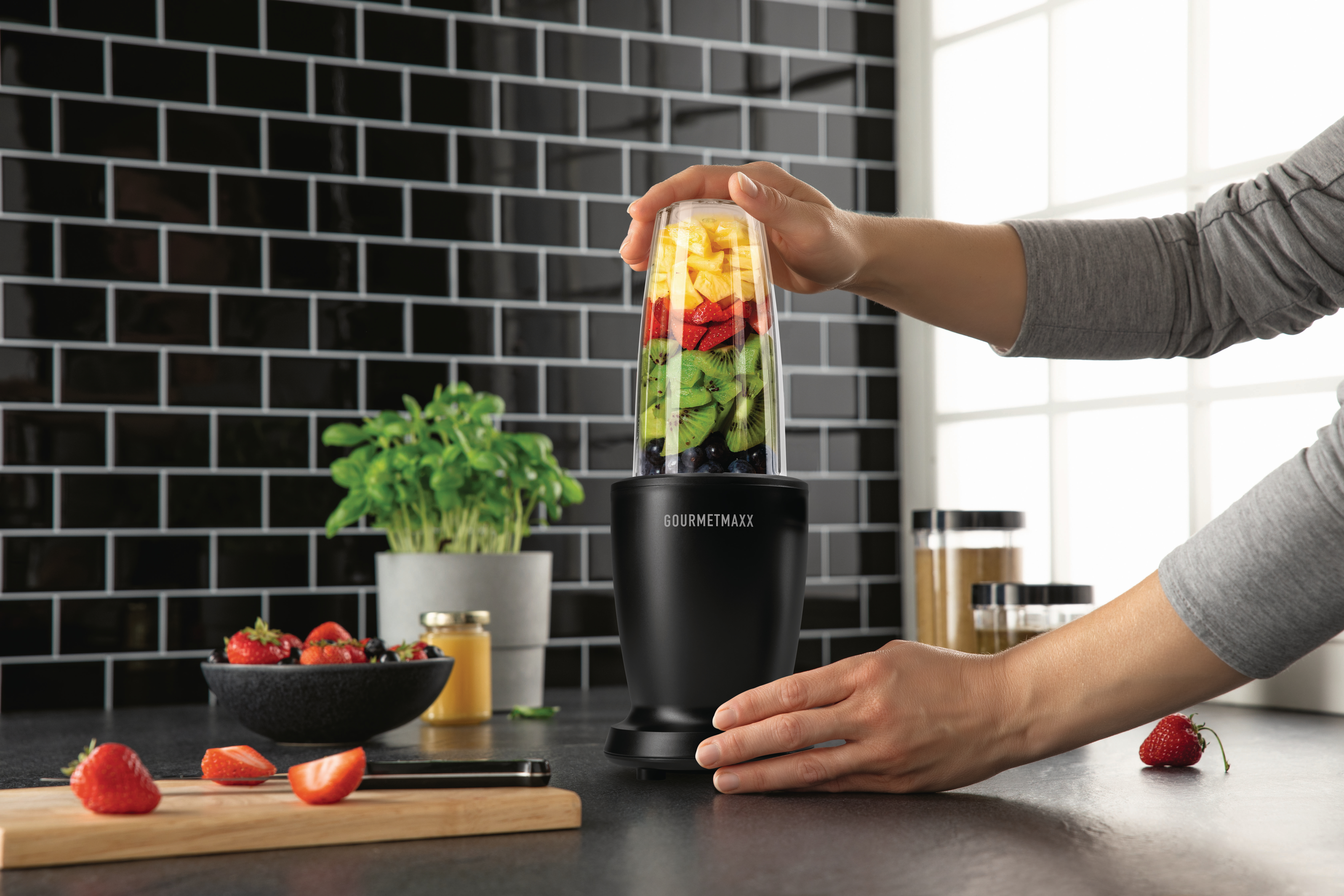 Eine Person bereitet mit einem Mixer einen Smoothie zu. Der Mixer ist mit Früchten gefüllt. Erdbeeren und eine Pflanze stehen auf der Theke.