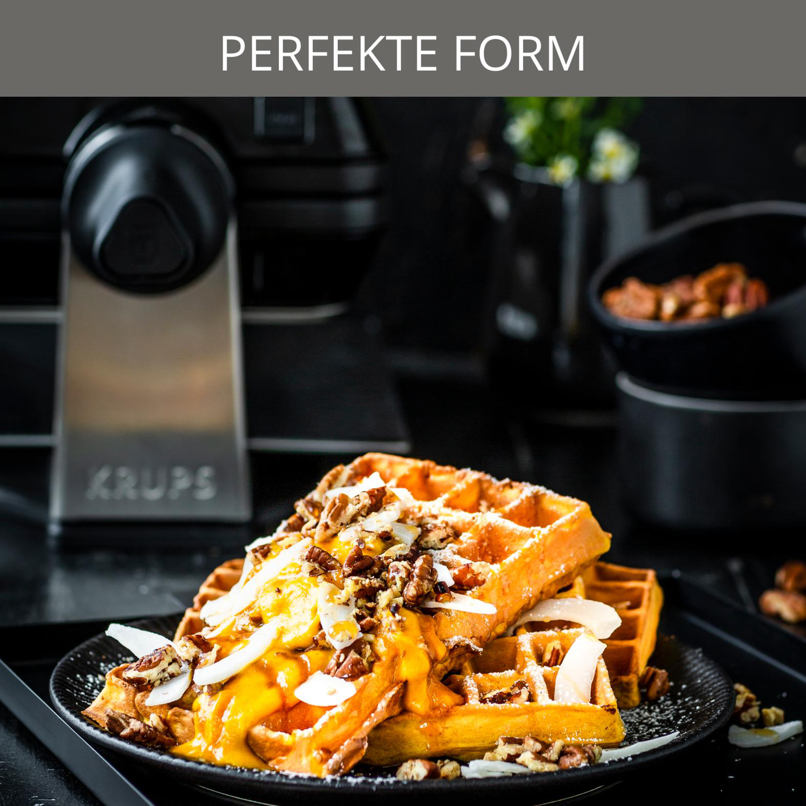 Waffeln mit Belag auf einem schwarzen Teller. Zutaten sind Soße, Nüsse und Kokosnuss. Im Hintergrund ein Krups Waffeleisen.