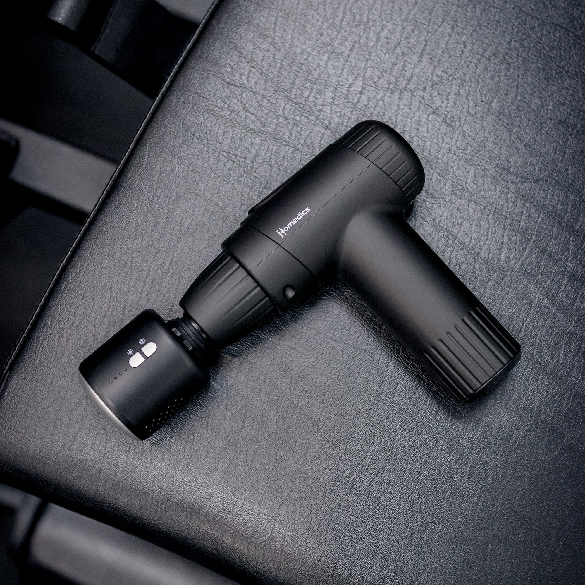 Czarny pistolet do masażu Homedics na czarnej skórzanej powierzchni, z wyposażeniem siłowni w tle.