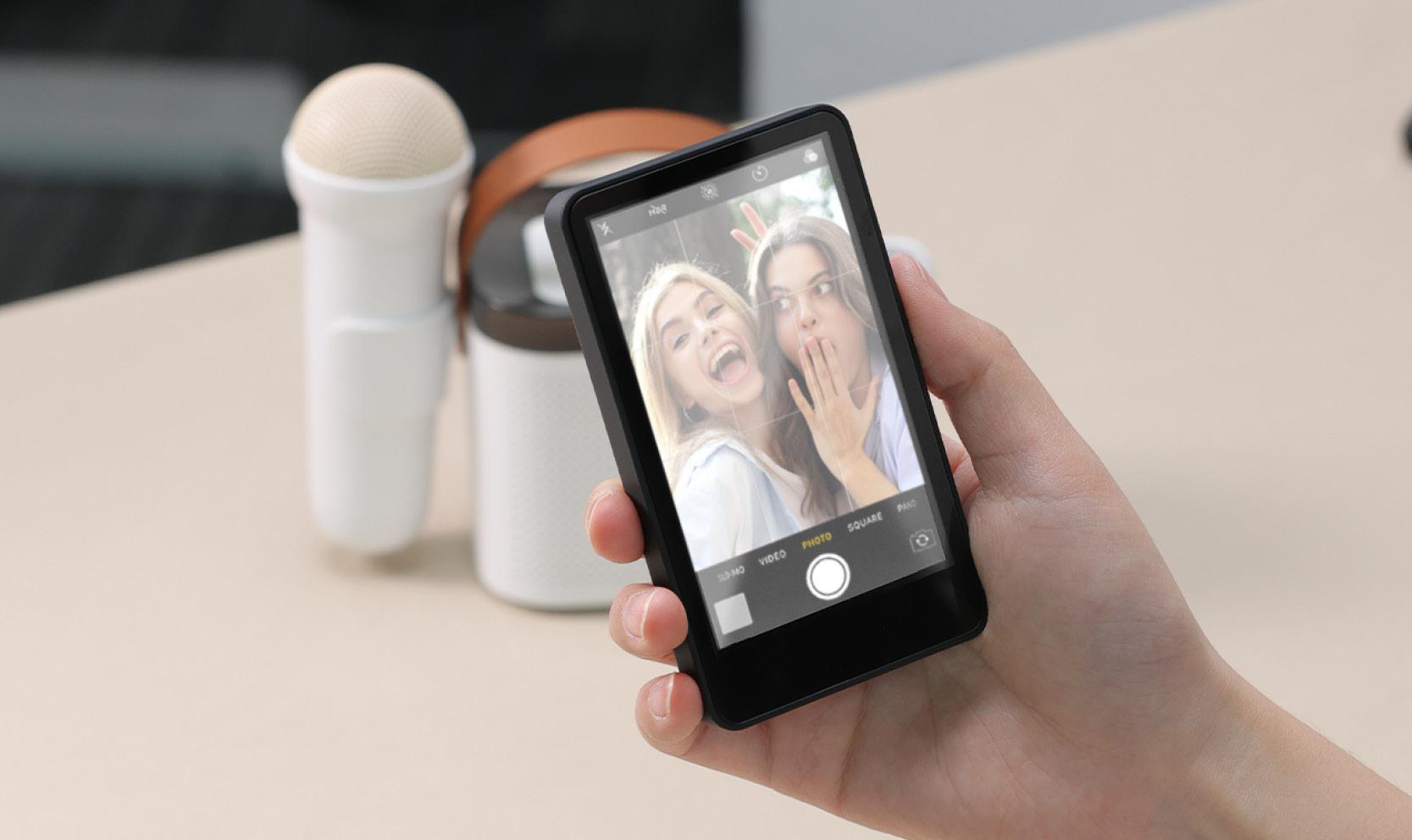 Hand hält ein Telefon, das ein Selfie von zwei Frauen zeigt, die Grimassen schneiden. Ein Mikrofon und ein Lautsprecher sind im Hintergrund.