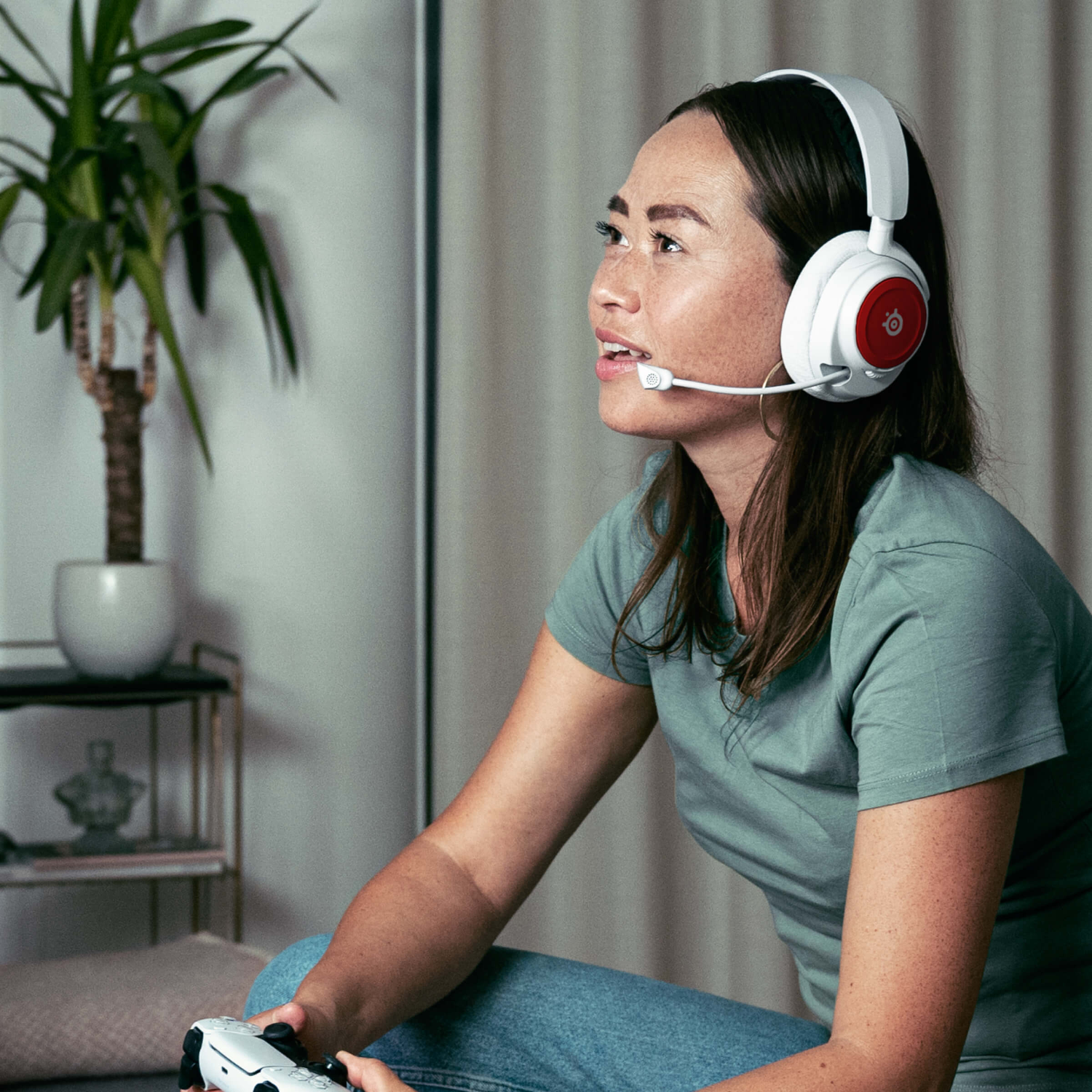 Woman wearing headphones and a mic, holding a game controller. She's looking up and to the right, with a plant and decor in the background.