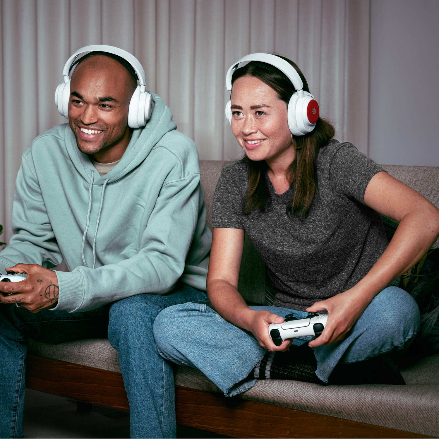 Two people on a couch wearing headphones play video games with controllers. They are smiling and focused.