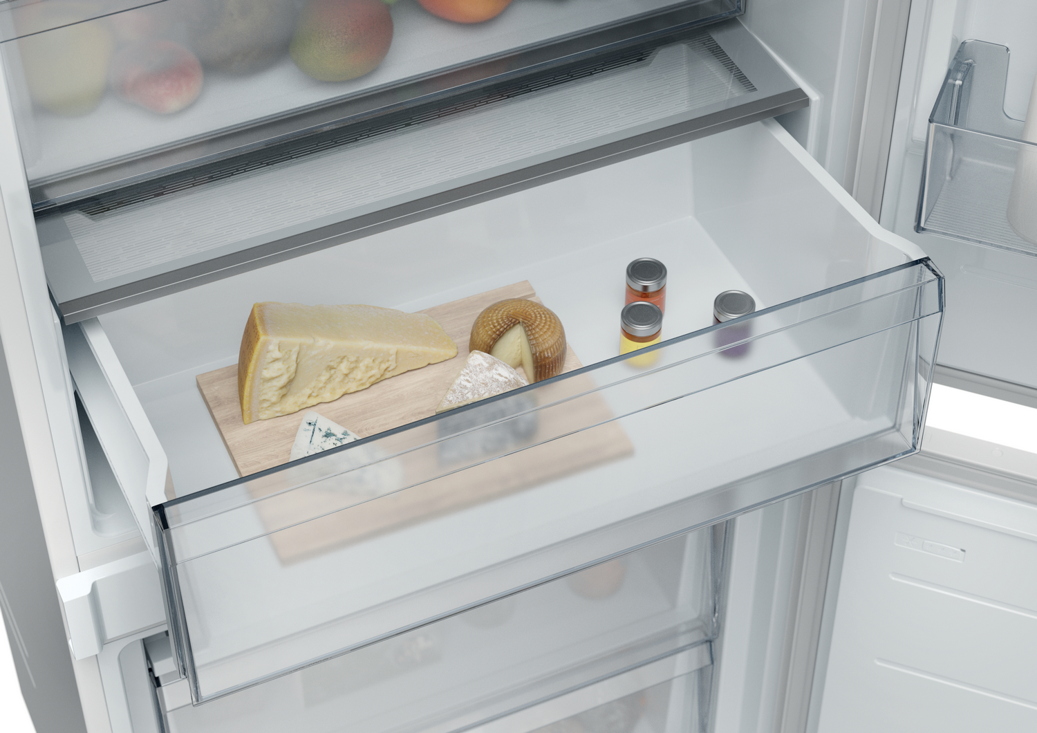 Cajón de refrigerador con varios quesos en una tabla de madera, pequeños frascos de condimentos y frutas arriba.
