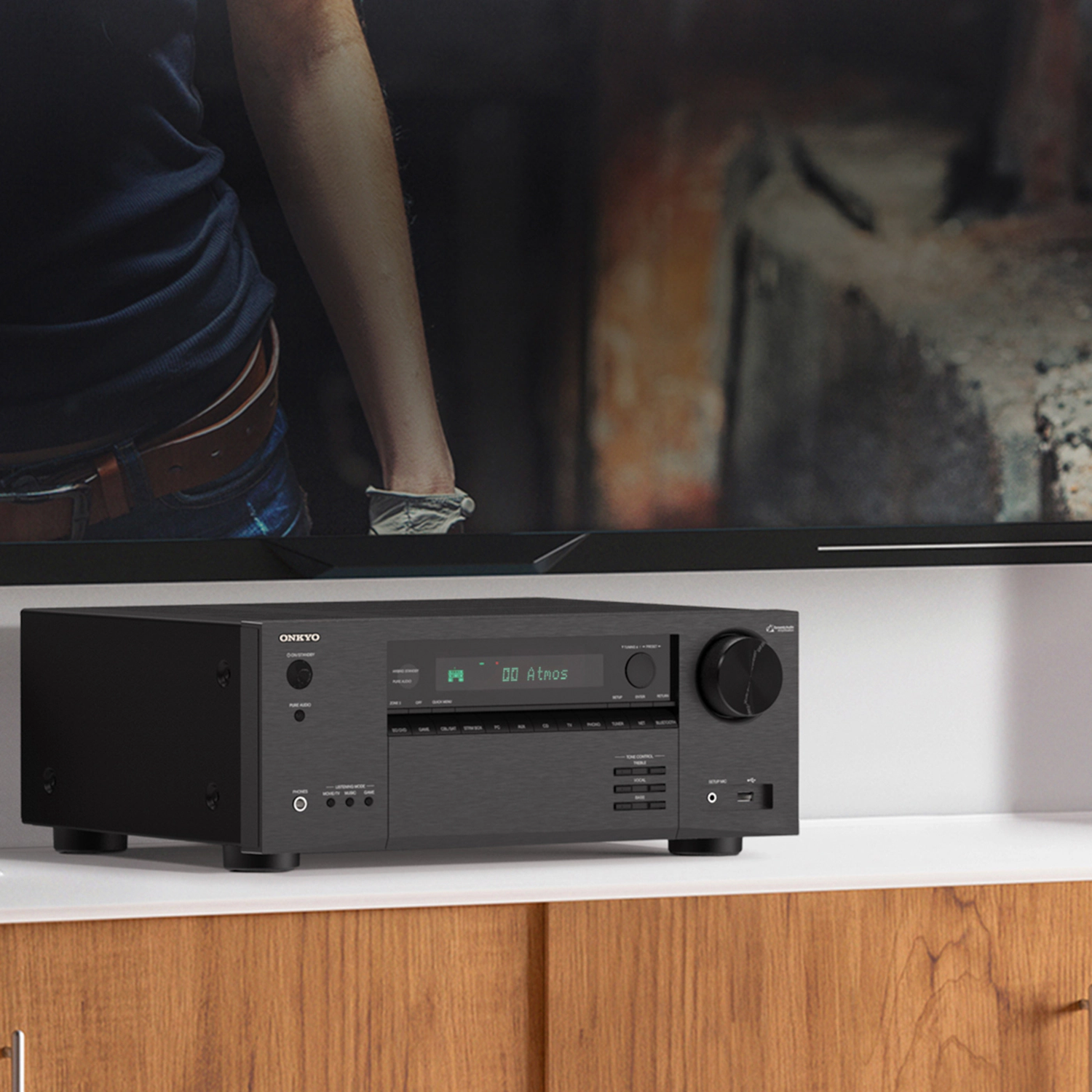 A black Onkyo receiver sits on a white shelf, below a TV. A person's arm and torso are visible in the background.