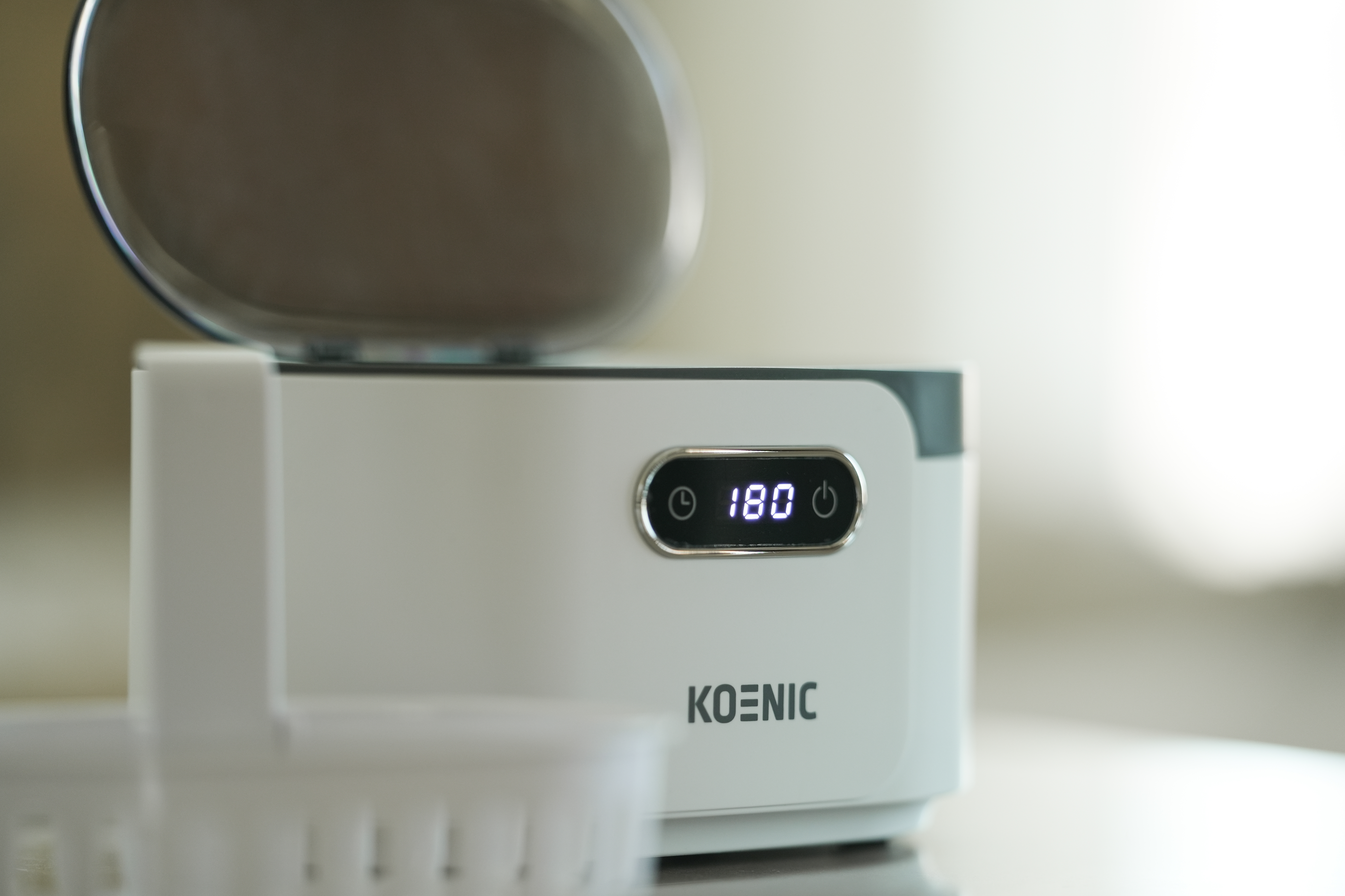 Un appareil Koenic blanc avec un couvercle ouvert affiche 180 sur un écran. Un objet blanc au premier plan.