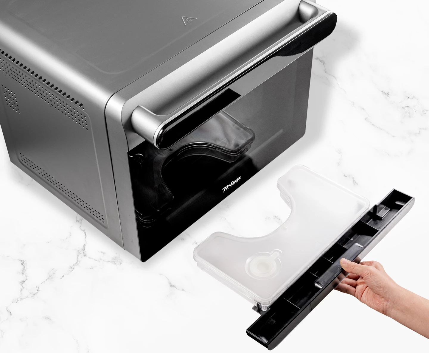 Four à micro-ondes argenté avec porte noire. Une main tient un récipient transparent et un plateau noir, sur une surface en marbre.