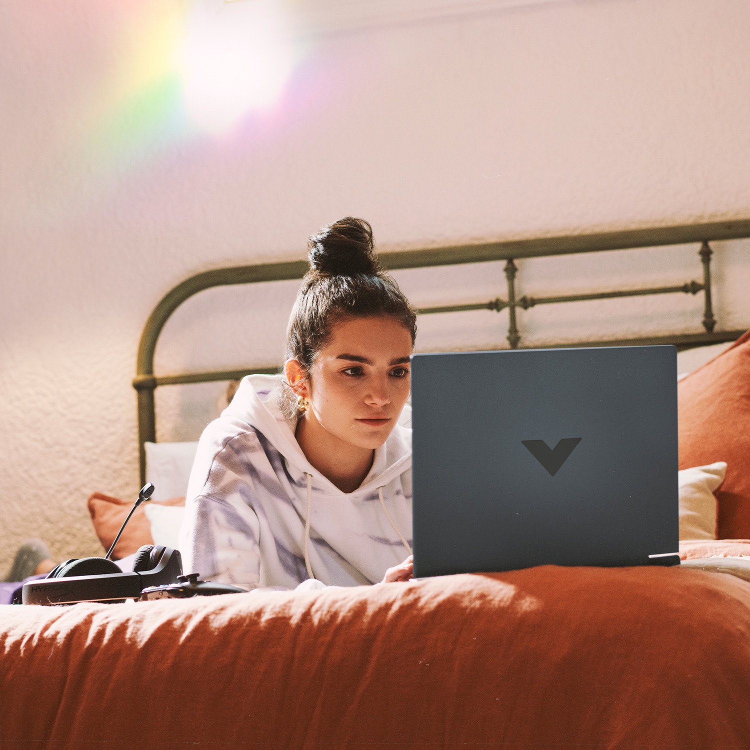A woman on a bed uses a laptop. She wears a hoodie, and there is a headset and controller nearby. The bed has orange bedding.