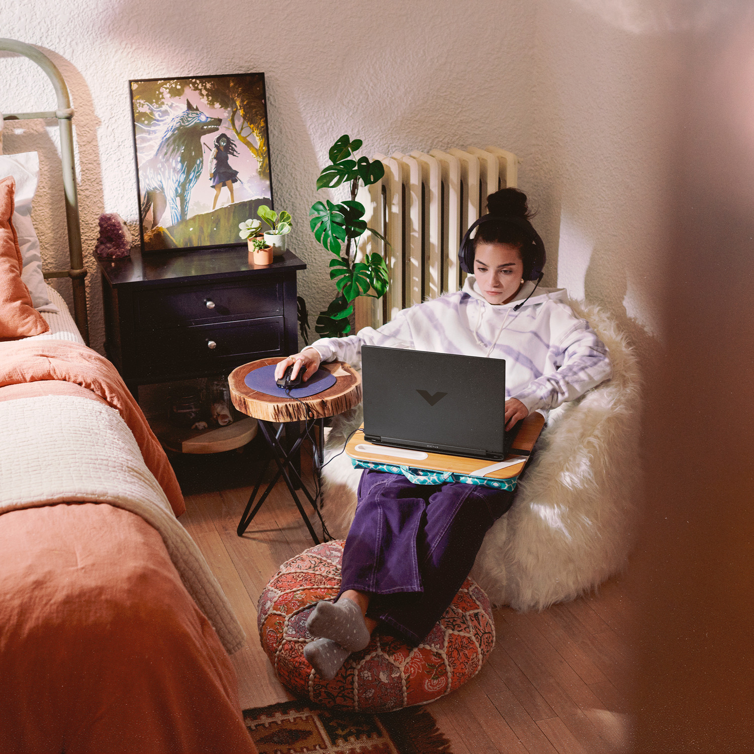 Woman in a fluffy chair using a laptop, headphones on. Bedroom setting with a painting, small table with mouse, and colorful ottoman.