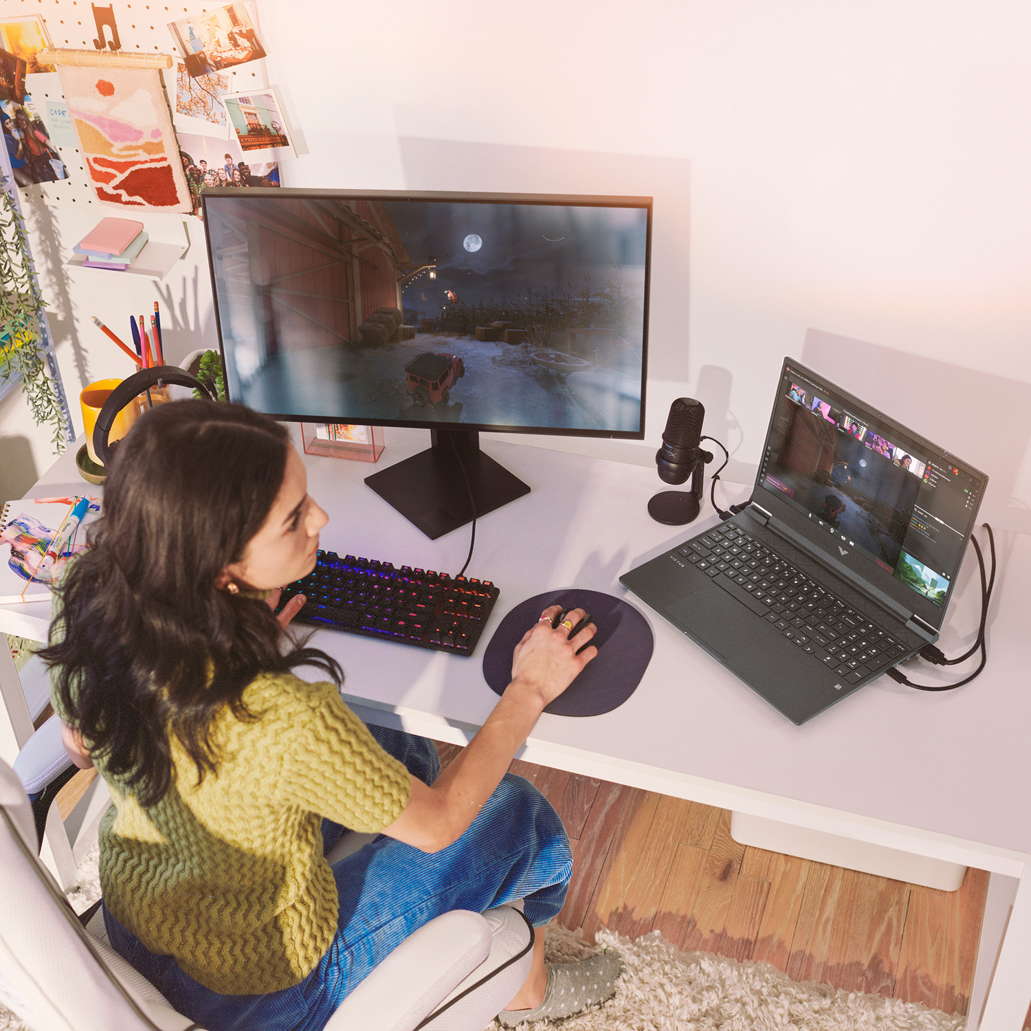 A woman sits at a desk gaming. She uses a computer, monitor, keyboard, and mouse. A microphone and laptop are also present.