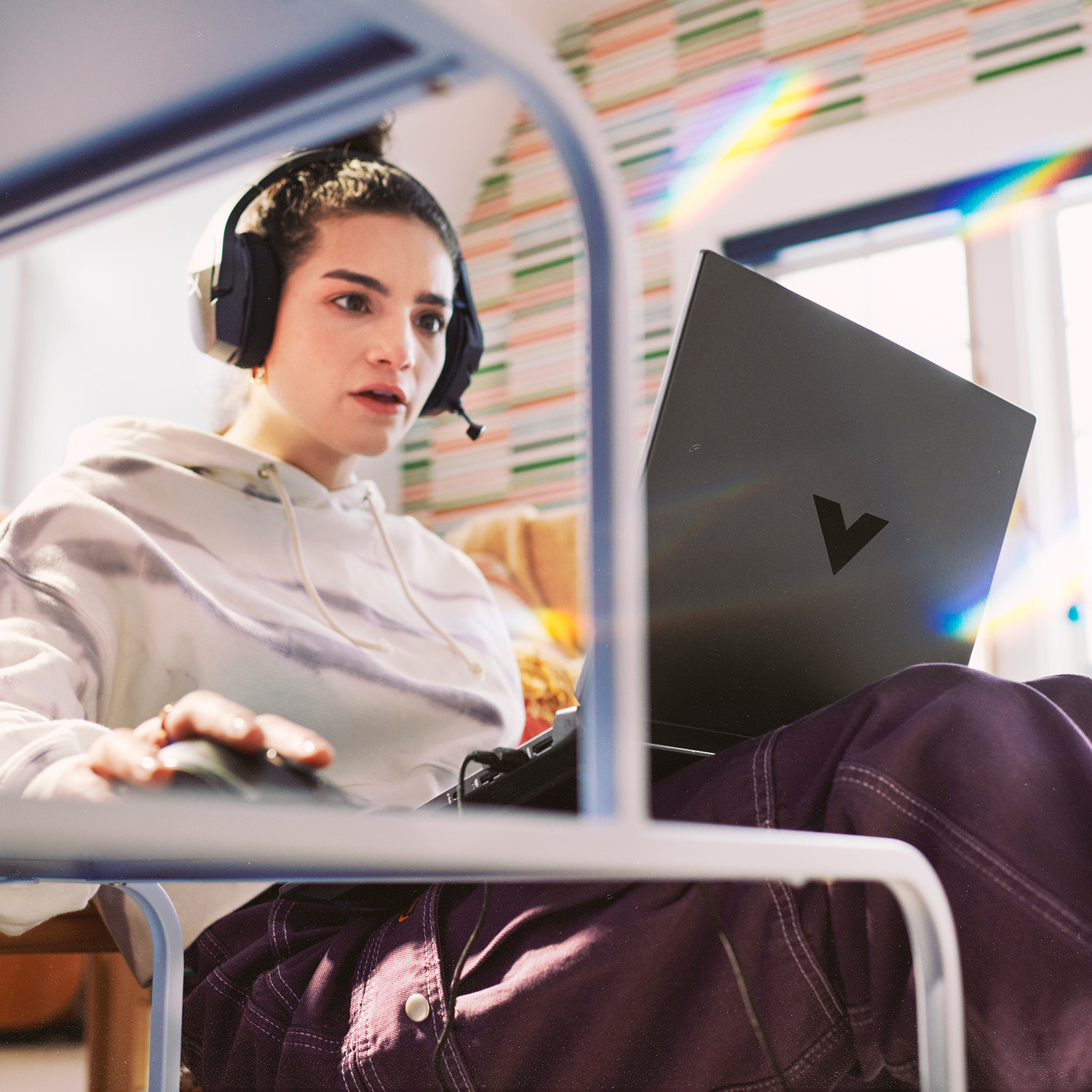 Woman wearing headset, using a laptop. She is seated at a desk. A rainbow is visible near a window in the background.