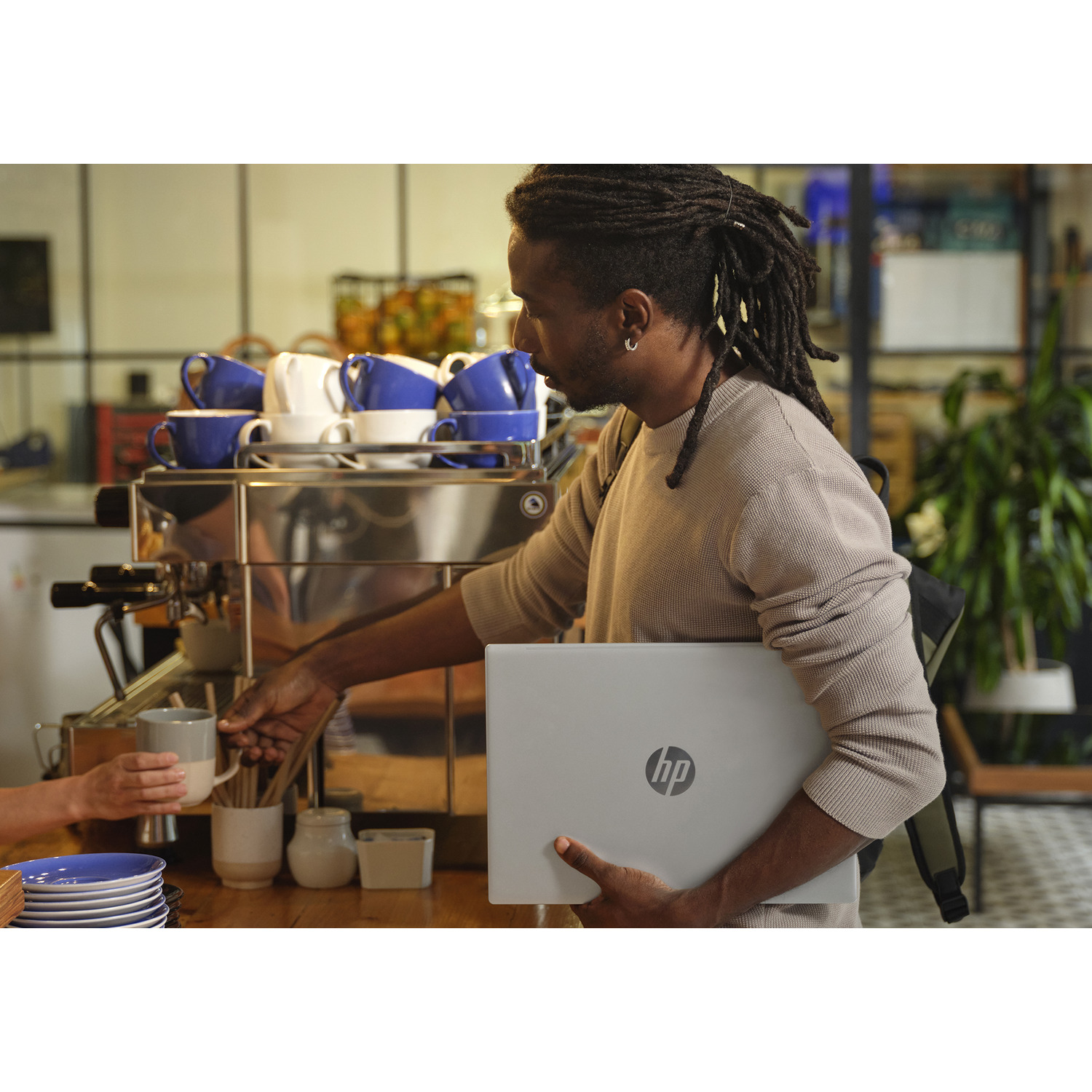 Man with dreadlocks holds a laptop, reaching for a cup at a coffee machine. A person hands him the cup. Background has cups.