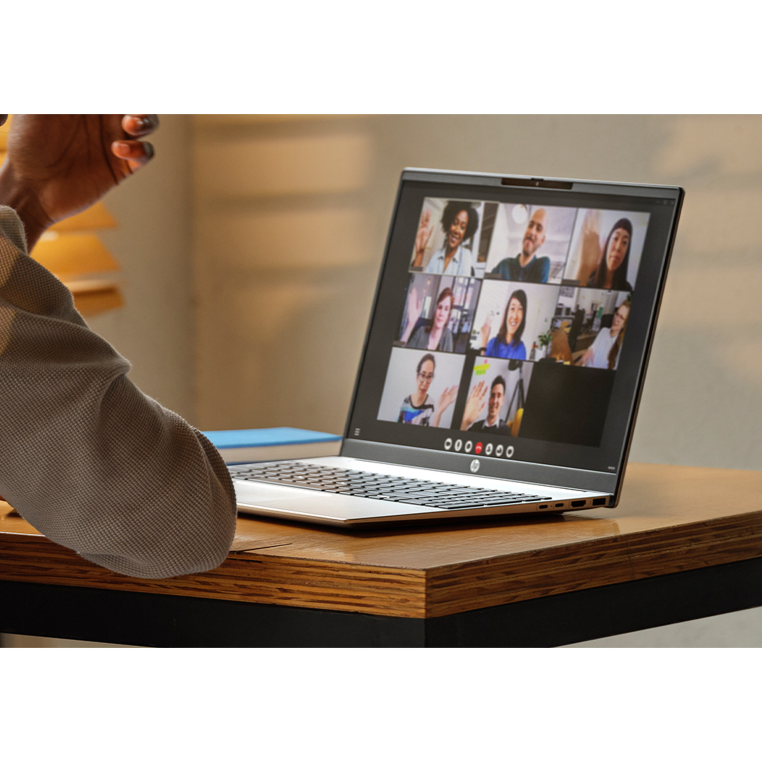 A person at a desk is on a video call. The laptop shows multiple people. The desk is wood. The person wears gray.