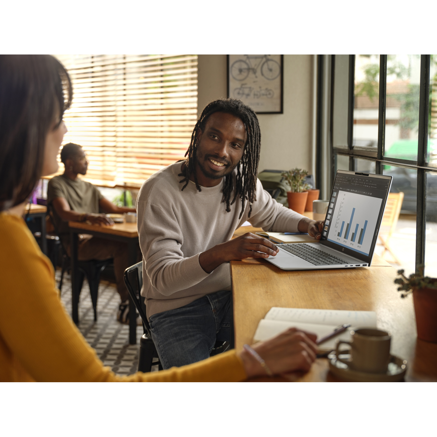 A man smiles at a woman while showing a laptop screen with graphs. Another person is in the background. A coffee cup is on the table.
