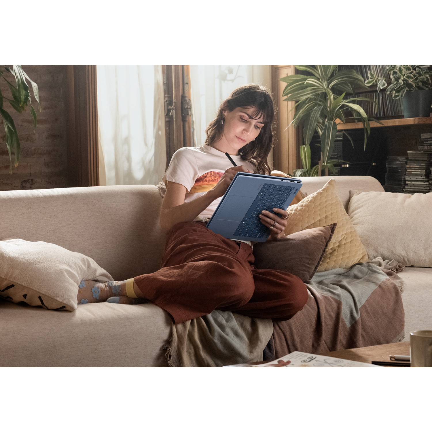 A woman sits on a sofa, writing on a tablet with a stylus. She wears a t-shirt and brown pants. Plants and books are in the background.