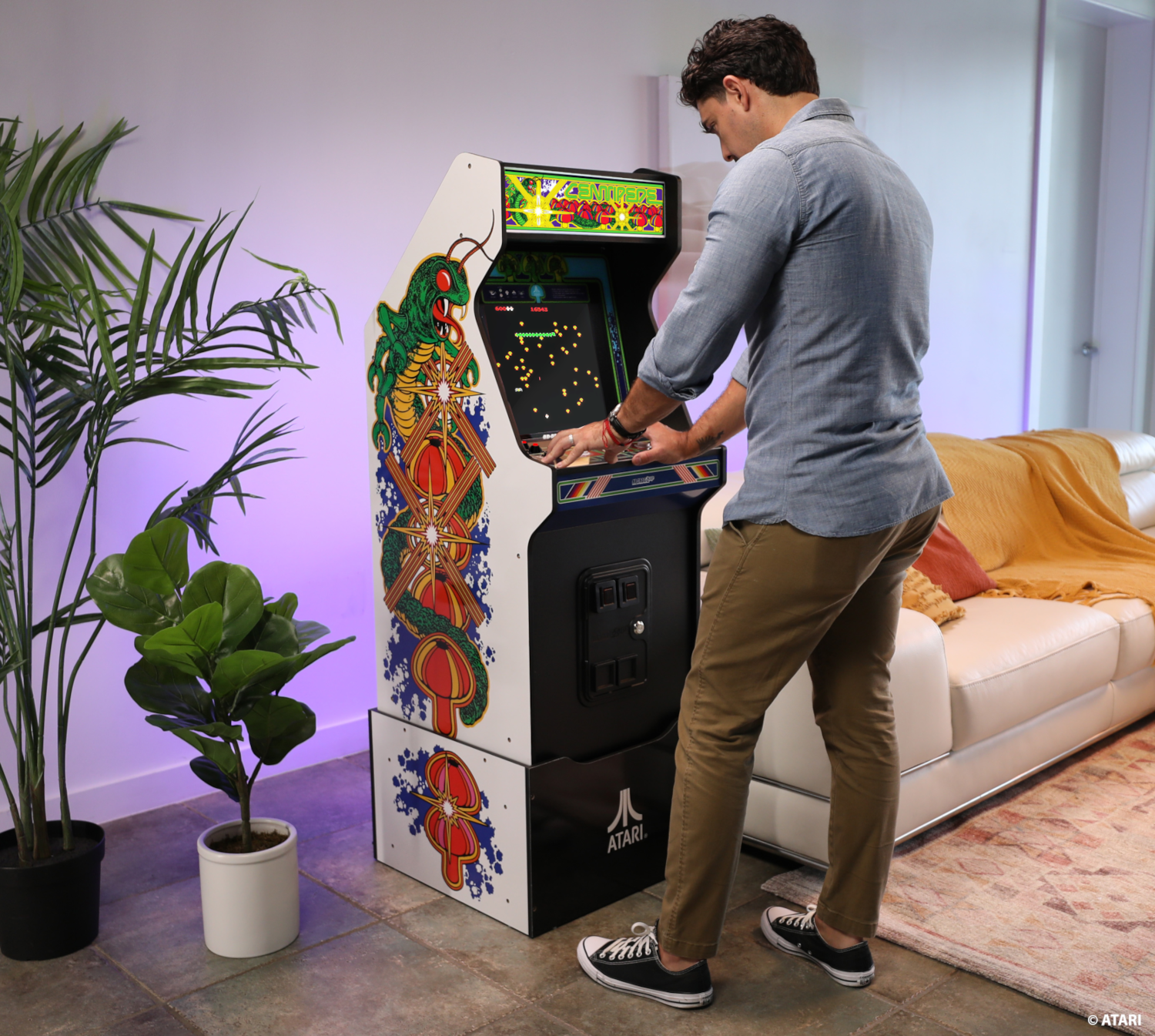 Egy férfi egy arcade-játékkal játszik. A gép színes, kígyós dizájnnal rendelkezik. Egy fehér kanapé és növények láthatók a háttérben.