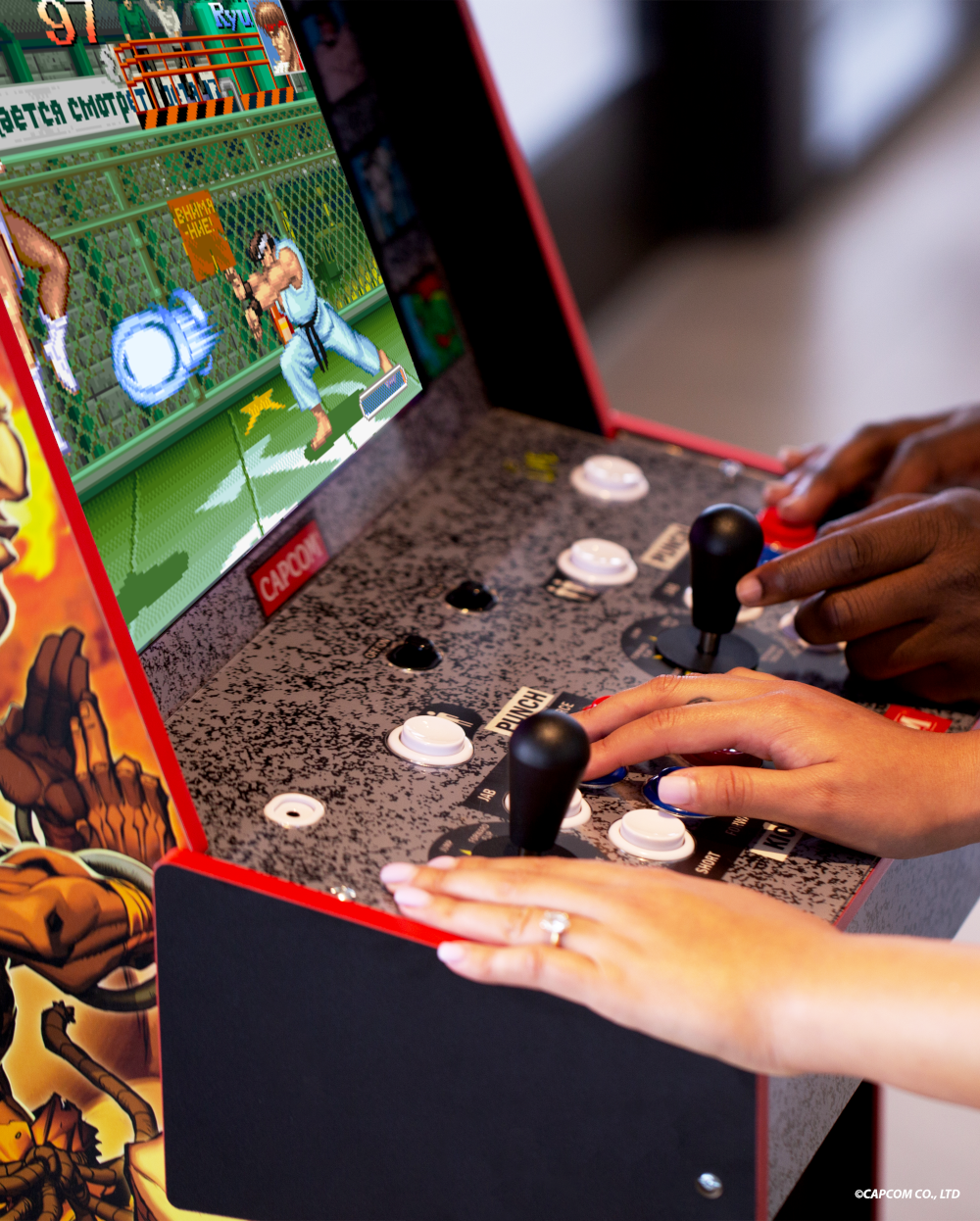 Kéz egy játéktermi játékkal játszik. A képernyőn egy verekedős játék látható. Gombok és joystick láthatók. Két ember játszik együtt.