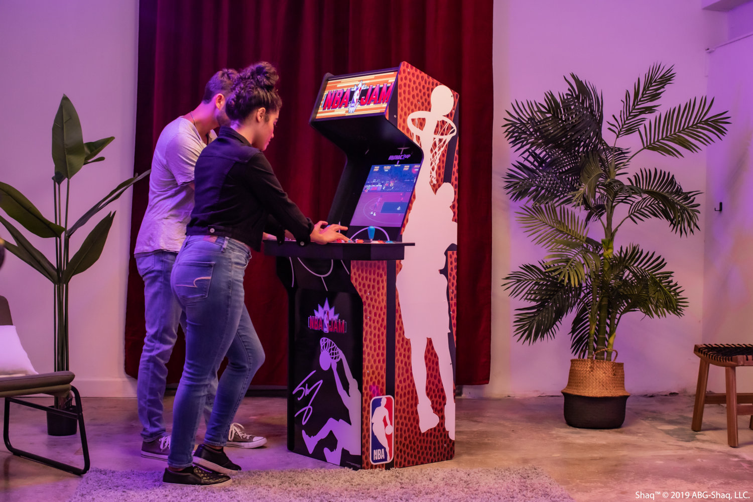 Két ember NBA Jam arcade játékkal játszik. Egy férfi és egy nő áll a gép mellett, kosárlabdapálya kijelzővel.
