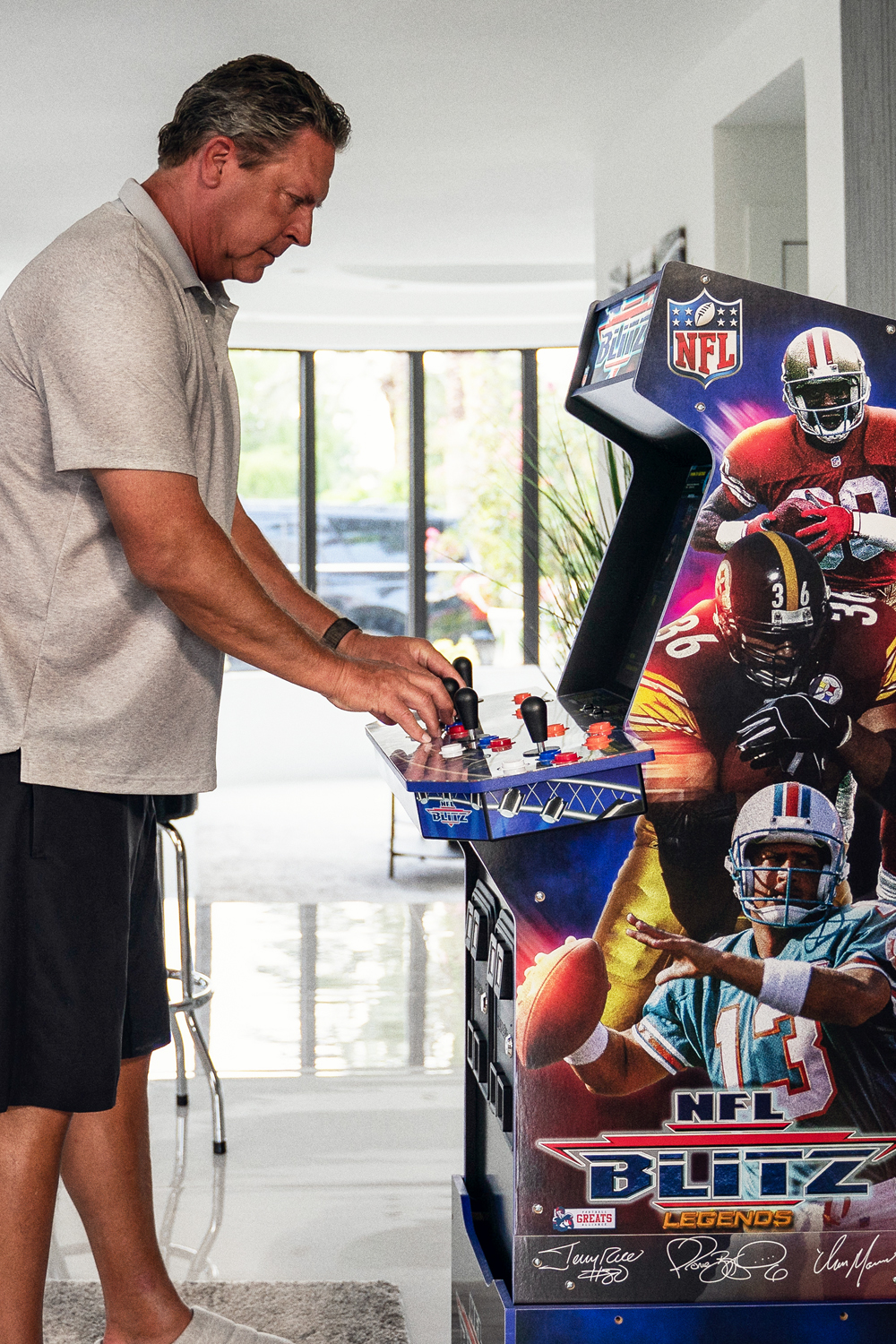 Egy férfi NFL Blitz arcade játékkal játszik. A gép elején NFL-játékosok láthatók. A férfi szürke inget és fekete rövidnadrágot visel.