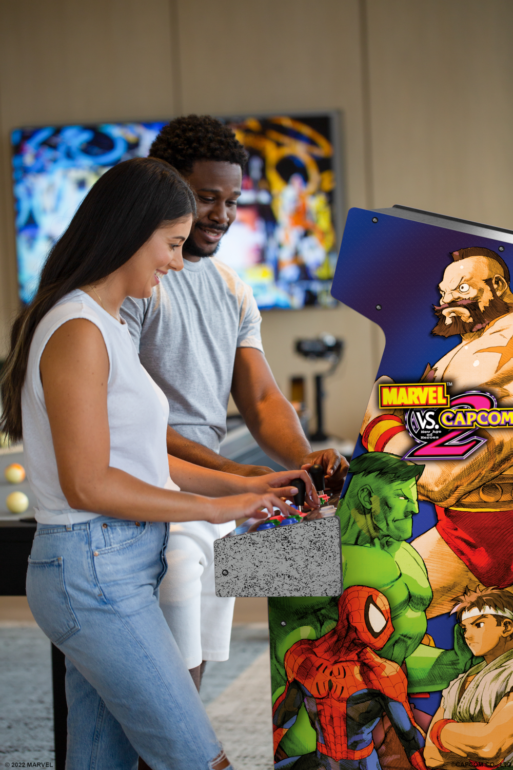 Egy pár játszik egy arcade játékkal. A gép Marvel vs. Capcom karaktereket jelenít meg, mint a Hulk és a Pókember. Háttérben egy TV.